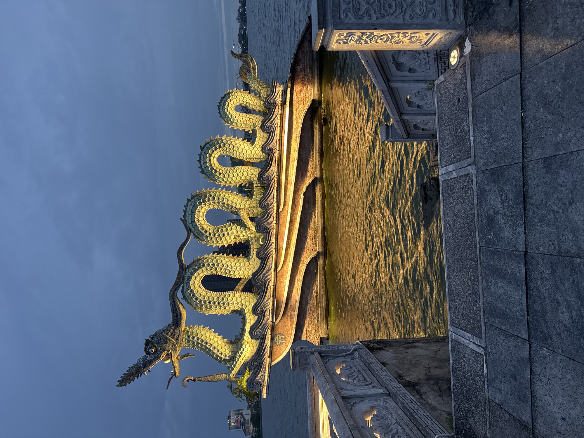 The image shows a large, illuminated dragon sculpture on a body of water, at dusk. The sculpture is positioned on a boat-like structure, with its body coiled and its head raised. The dragon is depicted with green scales and is lit from below with warm yellow lights, which reflect on the rippling water. In the background, the silhouette of a city skyline is visible, with several buildings. In the foreground, there are stone stairs and ornate railings, leading down towards the water and the dragon sculpture. The sky is a deep blue with some clouds, indicating it is late evening. This scene is located at Hồ Lục Thủy (Emerald Lake) in the heart of Hà Nội, Vietnam. There are no people visible in the image.