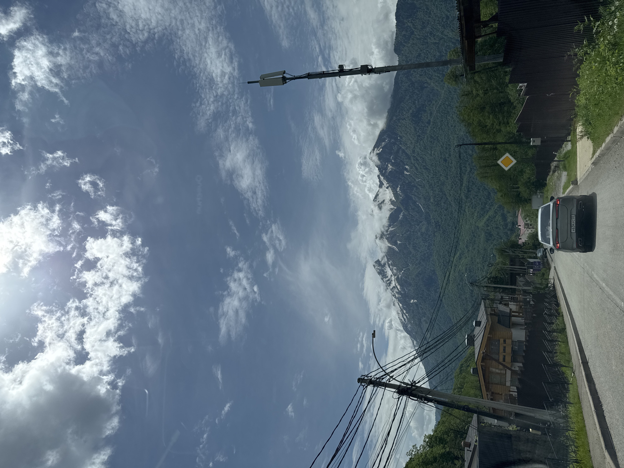 The image shows a road in Krasnaya Polyana, Russia, with a mountain range in the background. The sky is partly cloudy. Along the road, there are utility poles with wires, buildings, and a vehicle. A yellow diamond warning sign is visible near the side of the road. The mountain range is partially covered with green vegetation and some snow.