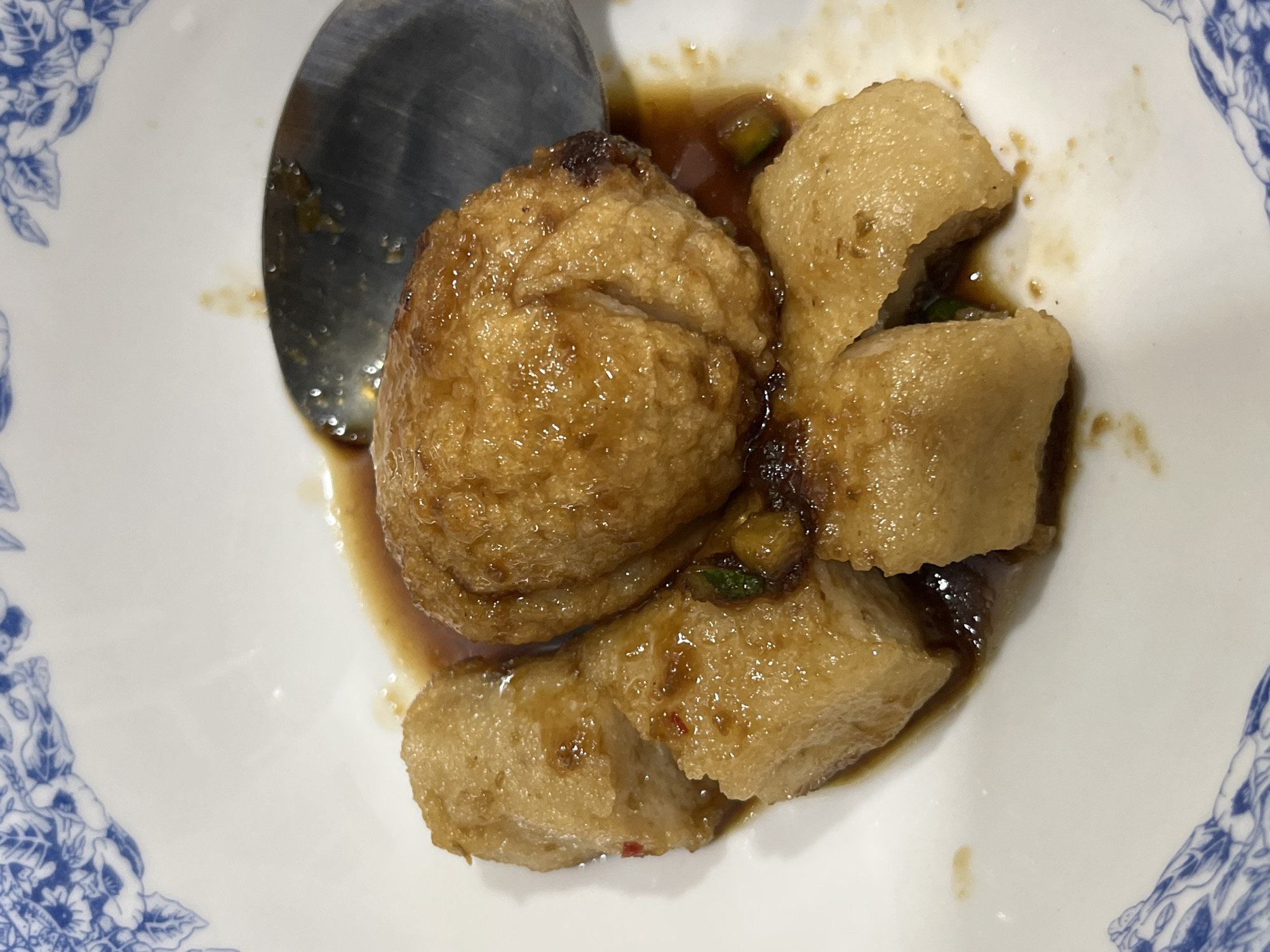 The image shows a close-up view of a dish containing what appears to be fried tofu or fish cakes served in a dark sauce. There are several pieces of the food, some cut into rectangular shapes and one larger, oval-shaped piece, all coated in the glossy sauce. A metal spoon is partially visible in the upper right quadrant of the image, resting in the sauce. The dish is presented in a white bowl with a blue floral pattern along the rim. There are no people visible in the image. The setting is likely a restaurant or home kitchen, given the presentation of the food. The image is not specific enough to determine a location in Teluk Air, Indonesia.