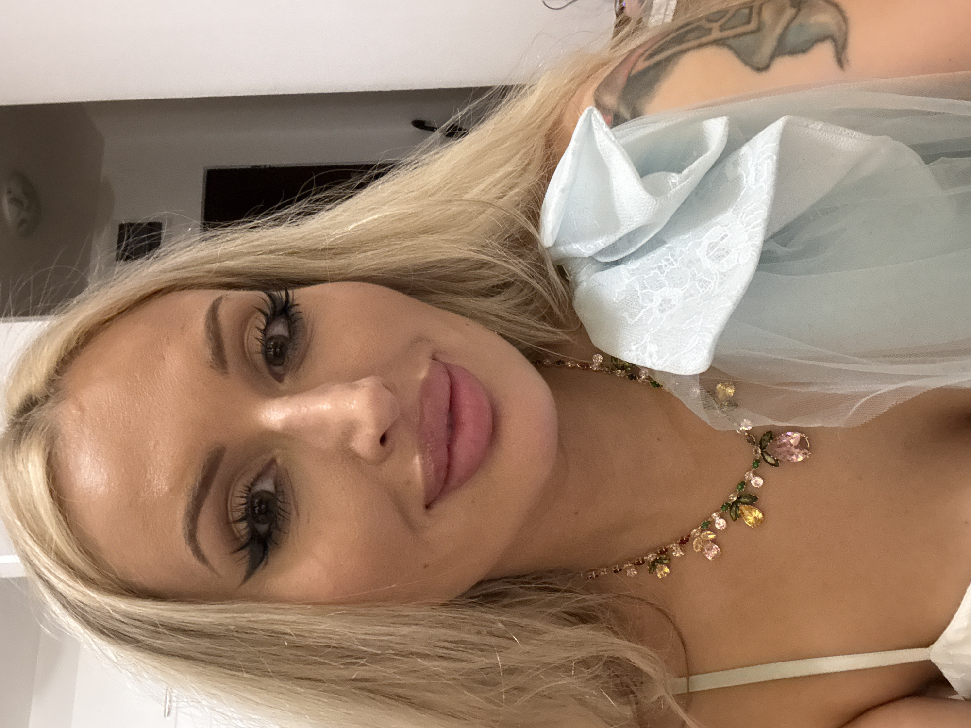 A close-up portrait, rotated counter-clockwise, features a young woman with long, straight blonde hair and fair skin, looking directly at the camera with a subtle smile. She has pronounced eye makeup, including dark eyeliner and long, full eyelashes, and her lips are full and appear glossy.
She wears a light-colored top with a thin strap visible on her right shoulder. A voluminous light blue fabric, possibly tulle or chiffon, with a delicate lace pattern and ruffled detail, drapes over her left shoulder. Adorning her neck is a statement necklace featuring an array of colorful, faceted gemstones in various shapes and sizes, including pink, yellow, and green stones, set on a thin chain.
Part of a tattoo, depicting a stylized wing or ornate design in muted tones, is visible on her upper right back/shoulder. The background is softly blurred, indicating an indoor setting with light-colored walls. The image focuses on her face, hair, and upper body, highlighting her jewelry and the intricate details of her attire. No text is visible.
