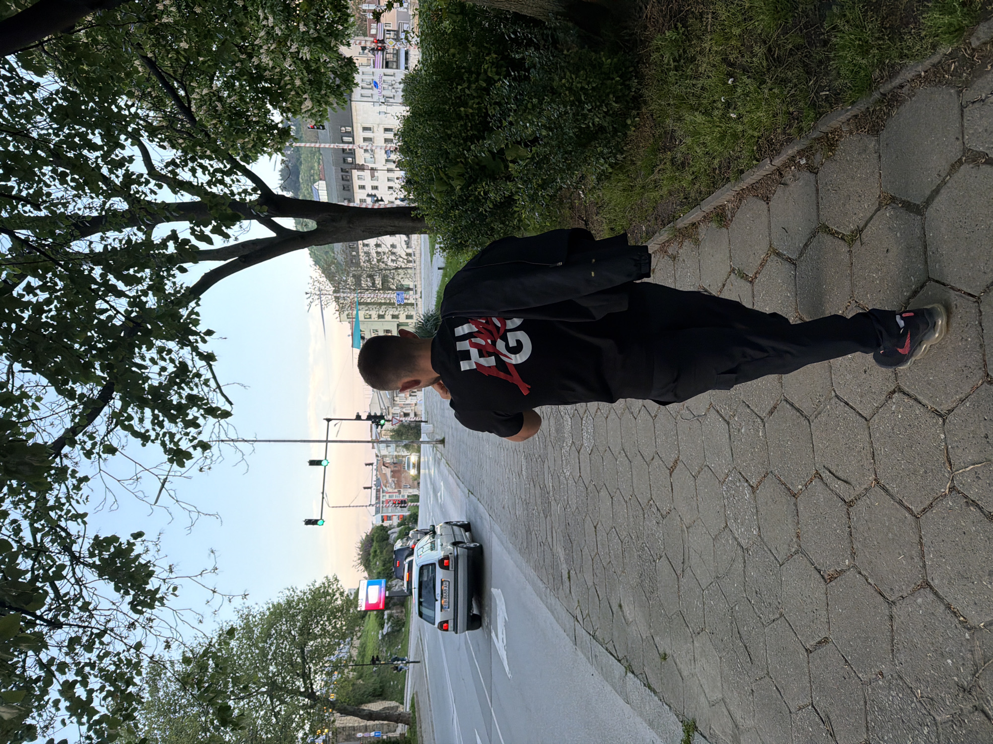 A man is walking on a sidewalk in Gabrovo, Bulgaria. He is wearing a black shirt with a white and red design on the back, black pants, and dark-colored sneakers. He is carrying a black jacket over his shoulder. The sidewalk is paved with hexagonal concrete blocks. A street runs parallel to the sidewalk, with cars driving on it. There are green traffic lights visible in the background. Buildings can be seen in the distance, as well as trees on both sides of the street. A billboard is also visible on the street. The overall scene appears to be a street in an urban setting in Gabrovo.