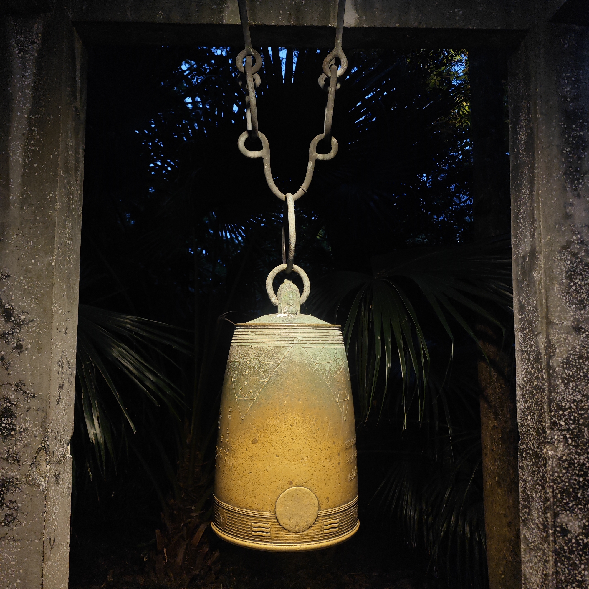 A bronze bell hangs from a chain between two concrete posts in Yuzhong District, China. The bell is tarnished, with geometric patterns at the top and a raised circle near the bottom. Palm trees and faint blue light are visible behind the bell. The posts are weathered.