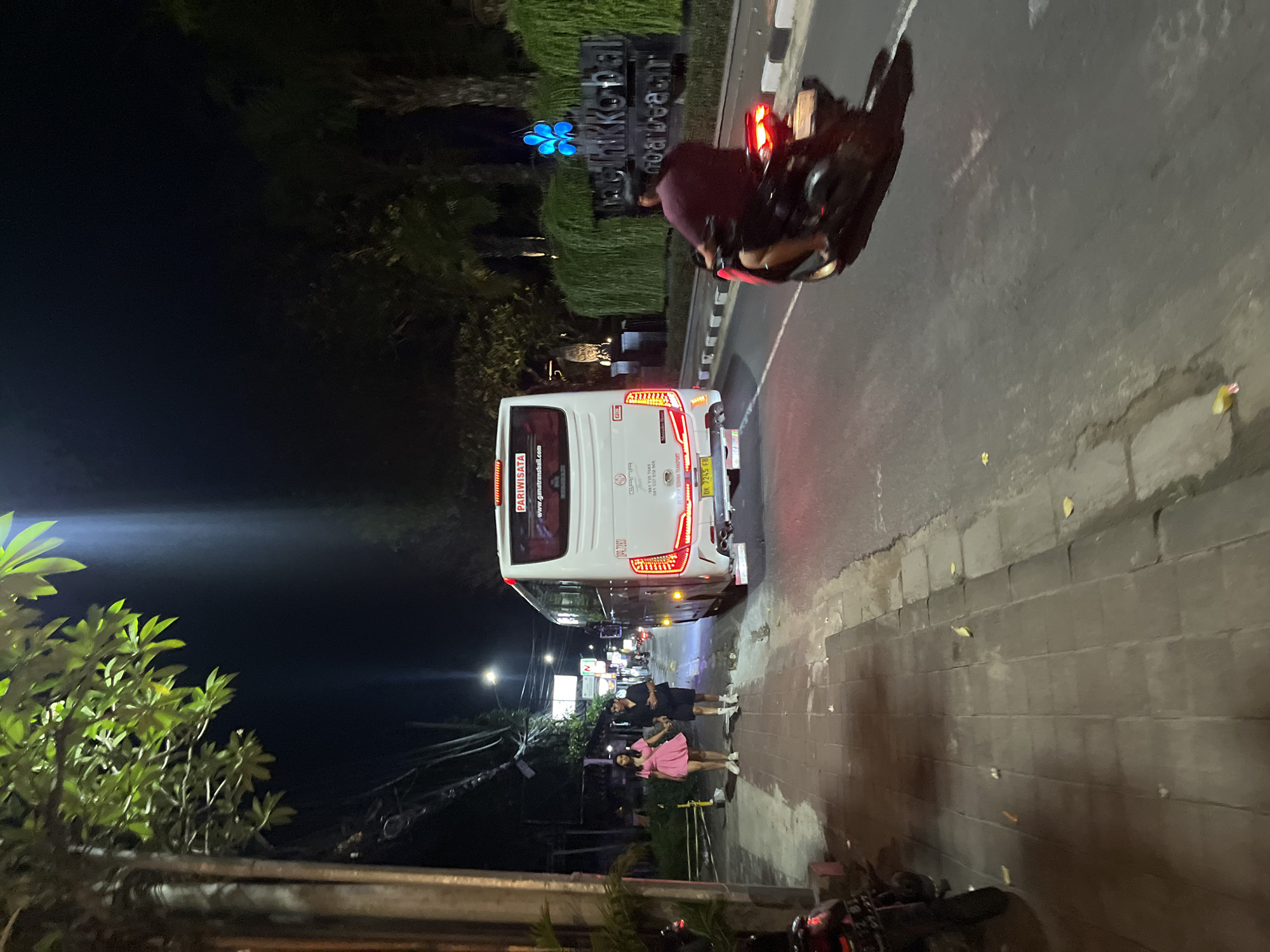 A nighttime scene in Kuta Selatan, Indonesia shows a white bus parked on the side of a road, with its taillights illuminated. Next to the bus, a man and a woman are standing. A motorcycle is also visible, parked near the sidewalk. Another motorcycle is driven along the road. Above the bus, a sign is lit with blue lights. The location is identified as "Thikko bal". Street lights are visible in the background, illuminating the scene.