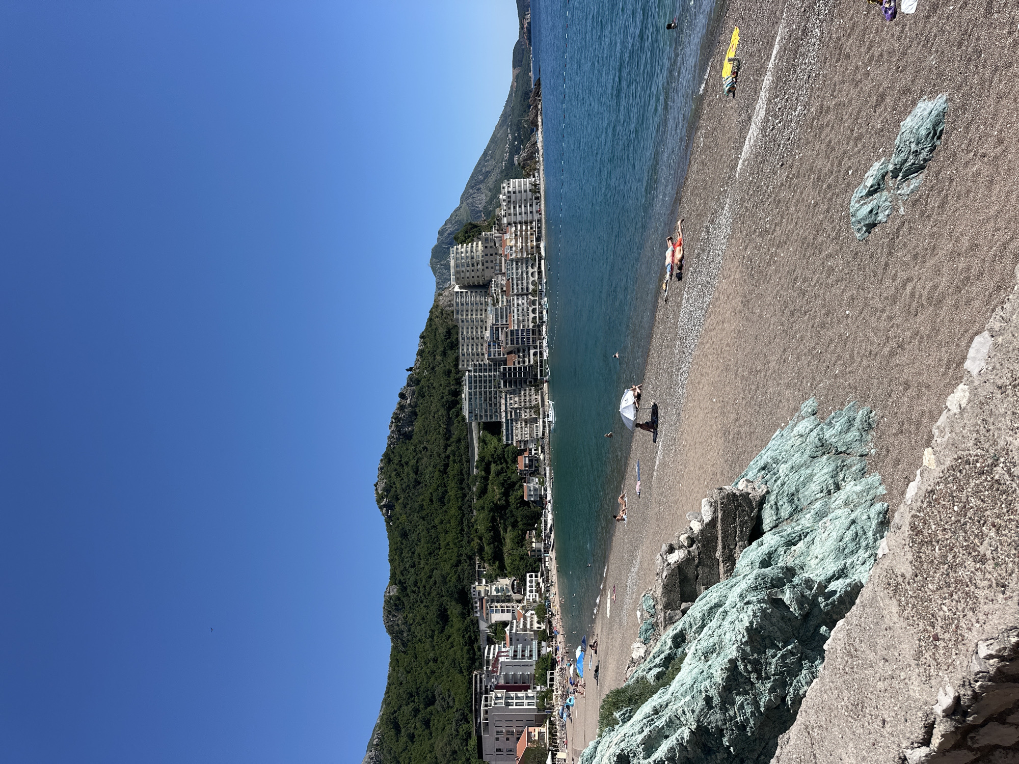 The image depicts a coastal scene in Rafailovići, Montenegro. The Adriatic Sea is on the right, transitioning to a pebble beach. Buildings line the shore, backed by a wooded mountain. A few people are on the beach, some under umbrellas, others swimming. Rocky formations with blue-green coloration are visible in the foreground. The sky is clear blue.