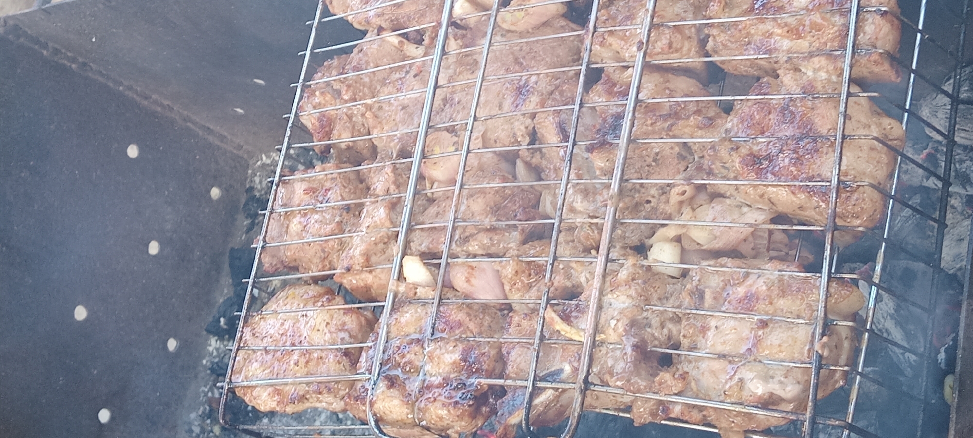 The image shows a close-up view of meat grilling on a barbecue rack. The meat appears to be thinly sliced and seasoned, with visible spices. It is arranged in multiple layers on a metal grill basket, which is placed over hot coals. Smoke is rising from the grill, indicating the cooking process is underway. The background suggests an outdoor setting, with the dark, textured surface of a grill pit visible to the left and bottom of the image. The lighting suggests it is daytime. The scene is devoid of people and text. The focus is entirely on the food being prepared, highlighting the texture and browning of the meat as it cooks.