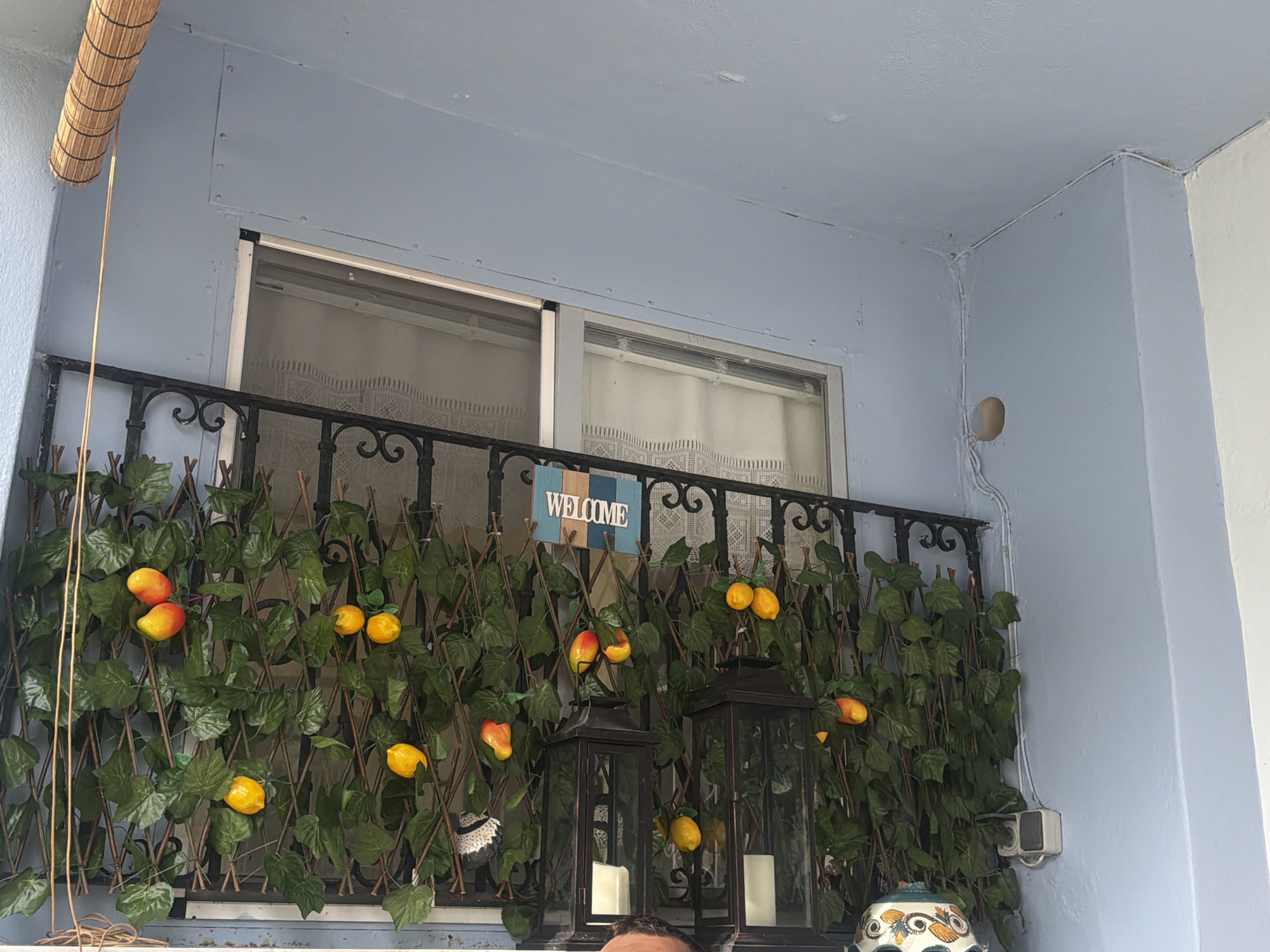 In Mandra, Greece, an outdoor balcony features a light blue ceiling and wall. A retractable bamboo shade is positioned in the upper left corner, with a thin cord extending downwards. The balcony railing is black and ornate. A trellis covered with artificial green leaves and faux lemons and oranges spans the length of the railing. A blue and white "WELCOME" sign hangs on the trellis. Two black lanterns, each containing a white candle, are placed on the balcony floor. A portion of a person's head is visible at the bottom center of the frame.