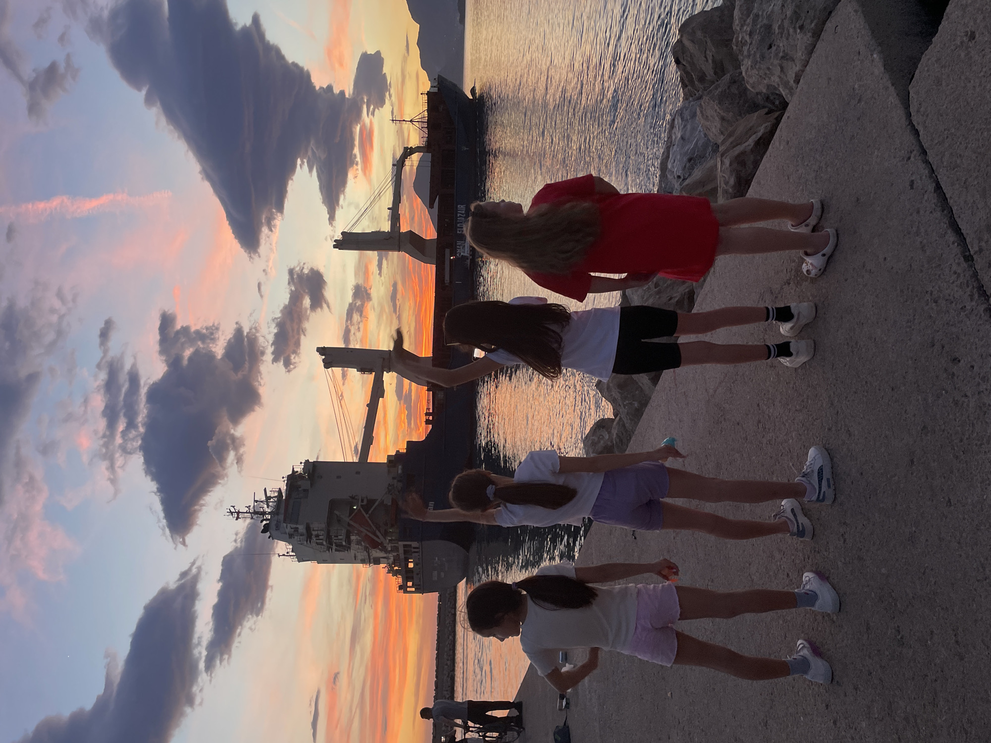 The image shows four young girls standing in a line on a concrete pier in Bar, Montenegro, silhouetted against a sunset sky and a large cargo ship docked in the harbor. The girls are of varying heights. The first one is dressed in a red dress, the second in a white shirt and black shorts, the third in a white shirt and lavender shorts, and the last one in a light yellow top and light pink shorts. They are positioned to appear as though they are touching or interacting with the cargo ship's cranes. The sunset paints the sky with orange, yellow, and purple hues. The cargo ship looms in the background. The pier is made of large concrete blocks, and some rocks are visible along the edge. In the bottom left, there are two men walking with a bicycle.