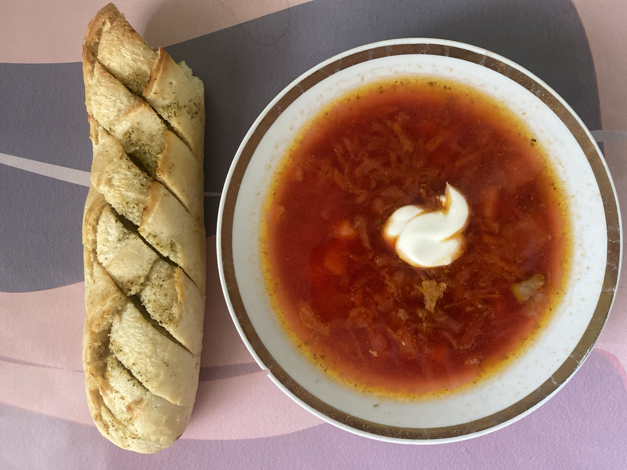 A plate of borscht, topped with a dollop of sour cream, sits next to a loaf of garlic bread on a table. The table appears to be indoors, potentially in a home. The background is a mix of gray and pink hues. No people are visible. The image does not inherently indicate the location being Chaikovsky, Russia.
