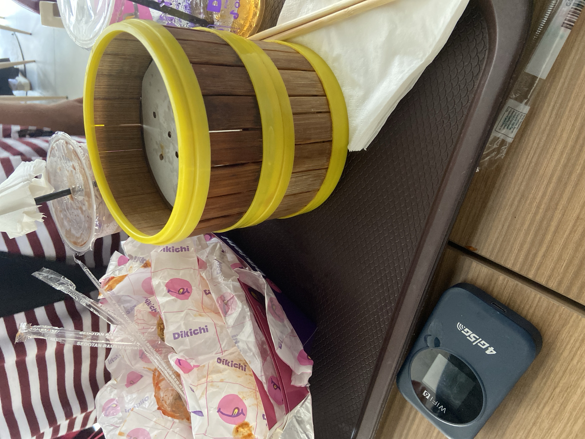 A close-up, eye-level shot shows a brown plastic tray on a wooden table with a variety of items for consumption. In the foreground, a dark blue portable WiFi device is positioned to the left of the tray, featuring a small screen and "4G 5G" branding. The tray contains a white paper napkin folded neatly, with two wooden chopsticks resting on top. To the right of the napkin is a bamboo-like container with yellow accents, filled with a white circular object, possibly food. Behind this, to the left, is a paper container filled with wrapped food items and clear plastic straws. Several clear plastic cups with drinks and lids are also visible in the background, one with a small white umbrella. A person wearing a maroon and white striped shirt is partially visible in the upper left corner. The scene appears to be set in Bago, Indonesia, likely at a cafe or restaurant.