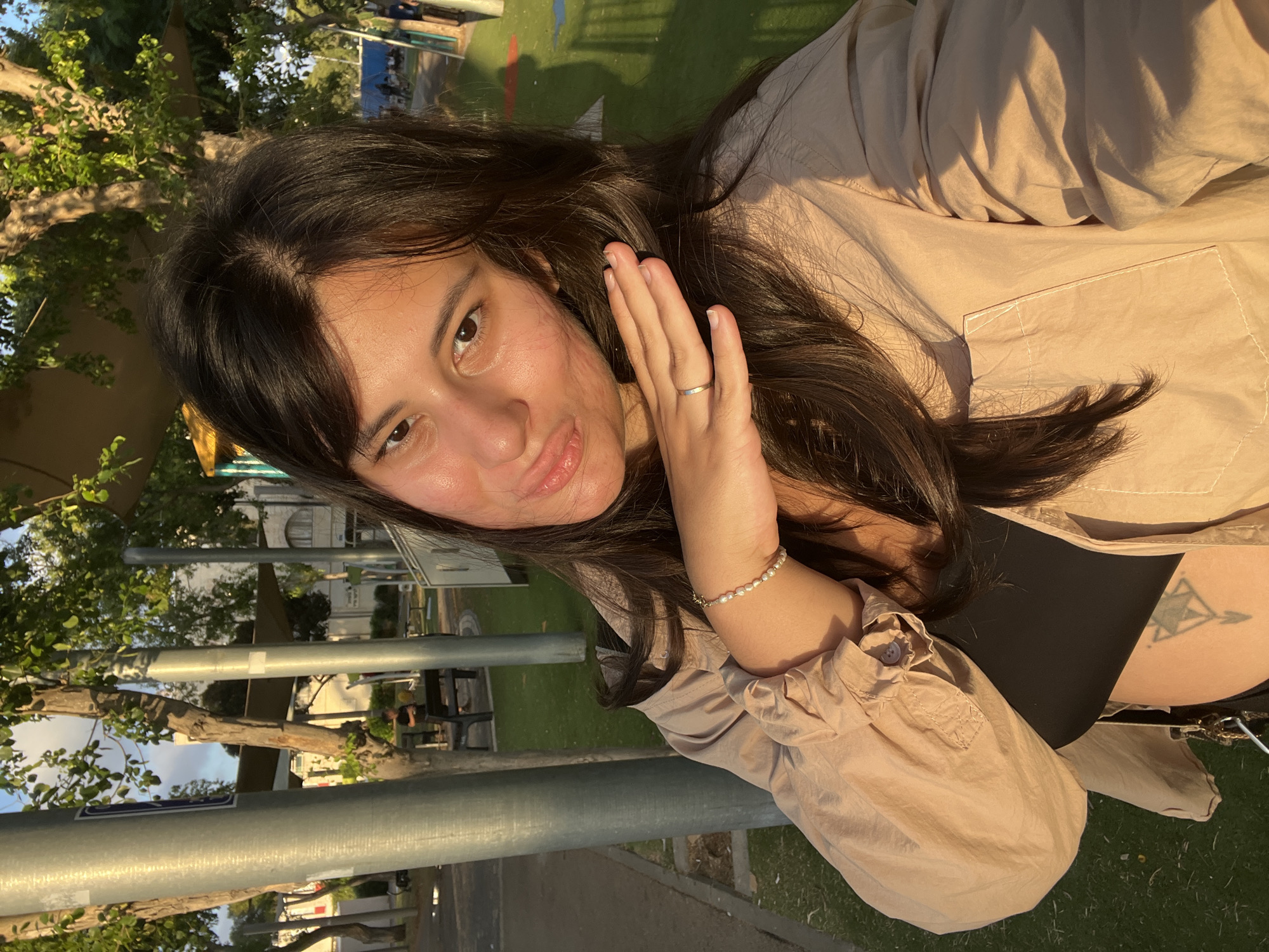 A young woman with dark brown hair is taking a selfie in an outdoor setting. She is wearing a light tan button-up shirt over a black top, and a pearl bracelet on her left wrist. A ring is visible on her left ring finger. Sunlight illuminates her face and hair, casting shadows on the ground. In the background, there are trees, a playground with green turf, and some buildings. The scene appears to be in a public park or recreational area, consistent with locations found in Kiryat Bialik, Israel. A partial view of a geometric tattoo is visible on her midriff.