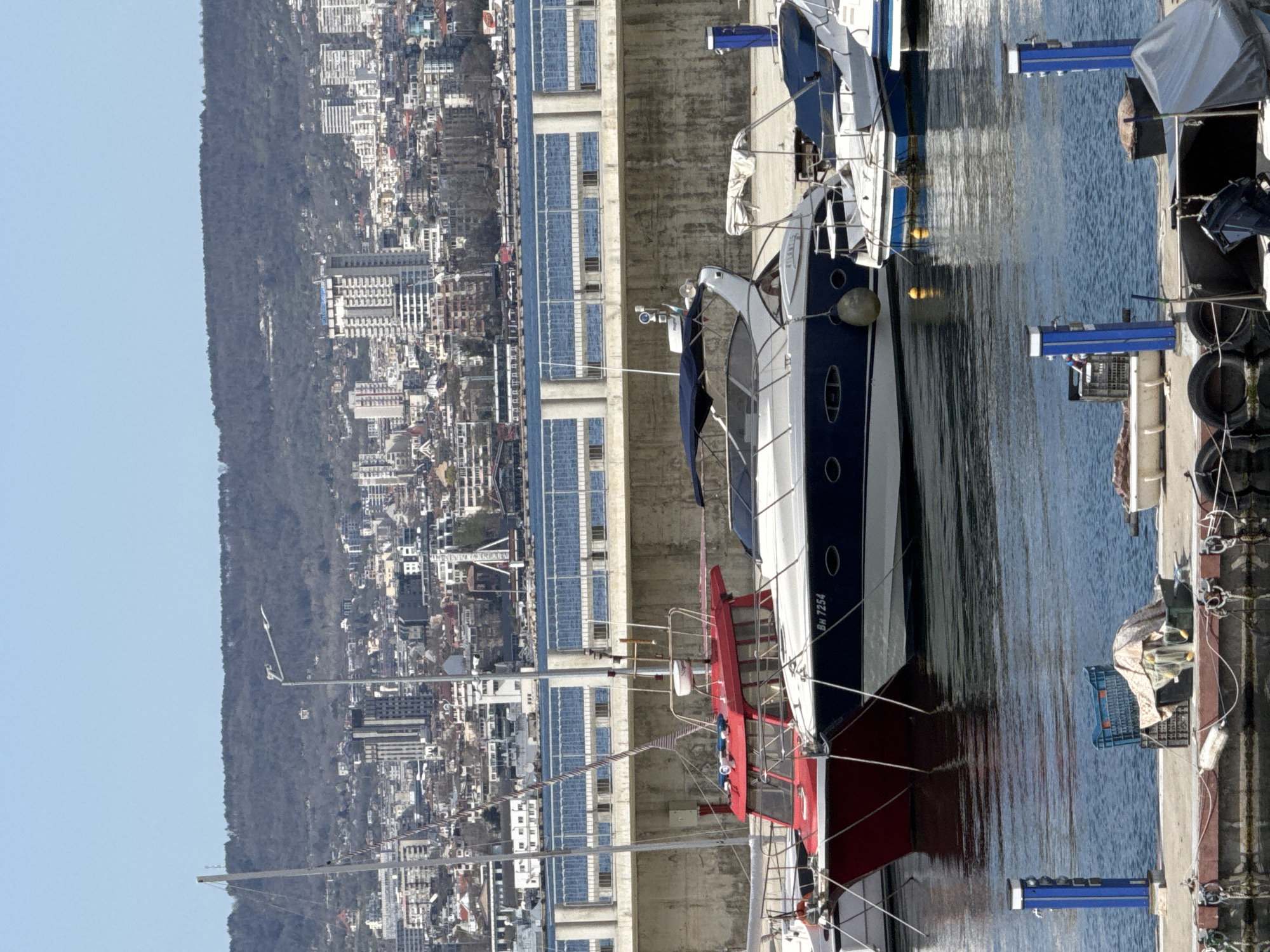 The image, oriented correctly, displays a sunny daytime scene of a marina in Varna, Bulgaria, with a city and a forested hillside in the background.
In the foreground, several boats are moored along a concrete pier on the right. The most prominent vessel is a white and blue motor yacht, featuring the registration "BH 7254" visible on its stern. Part of a red and white boat is also visible in front of it. The calm water of the marina reflects the boats and the clear blue sky. Blue service pedestals, likely providing power and water, are spaced along the pier. Mooring ropes secure the boats to the dock.
The background reveals a bustling city built into the side of a large, dark green, densely forested hill. Numerous buildings of various sizes and architectural styles, including multi-story structures, are visible, extending from the waterfront up the hillside. The sky above is clear and light blue, suggesting fair weather.
No people are clearly visible, and no specific activity or event is taking place beyond the general scene of a functioning marina. The setting conveys a sense of urban coastal life.