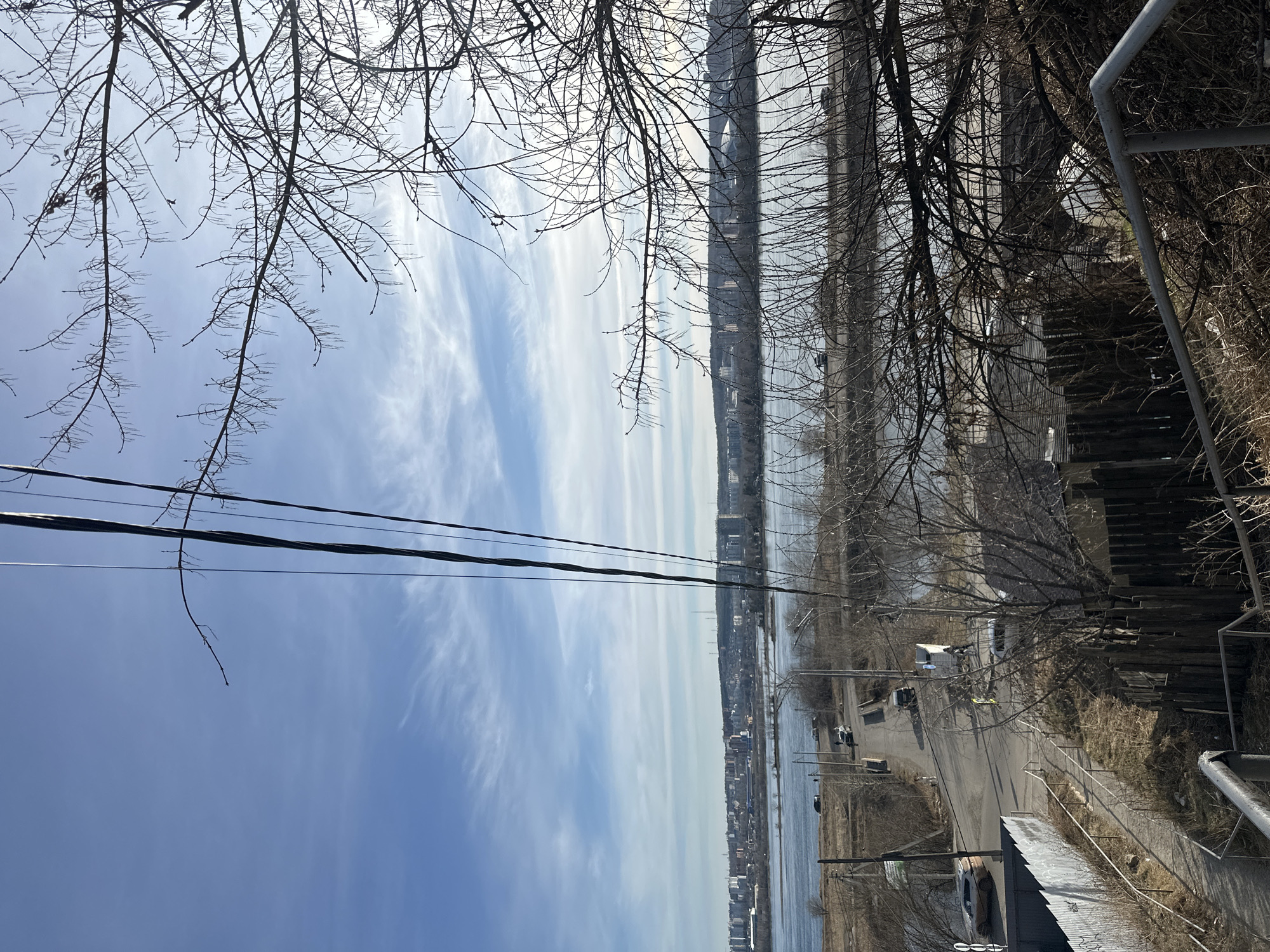 An oblique, eye-level shot looks over a city road and river under a blue sky streaked with clouds. Bare tree branches frame the top and right side of the frame, and power lines cut across the left side of the sky. A staircase with a gray metal railing runs along the right edge. The road has several parked vehicles and connects to a larger river. Across the river are buildings, suggesting a city skyline, possibly the cityscape of Irkutsk, Russia. The general landscape hints at a transition between urban and natural environments.