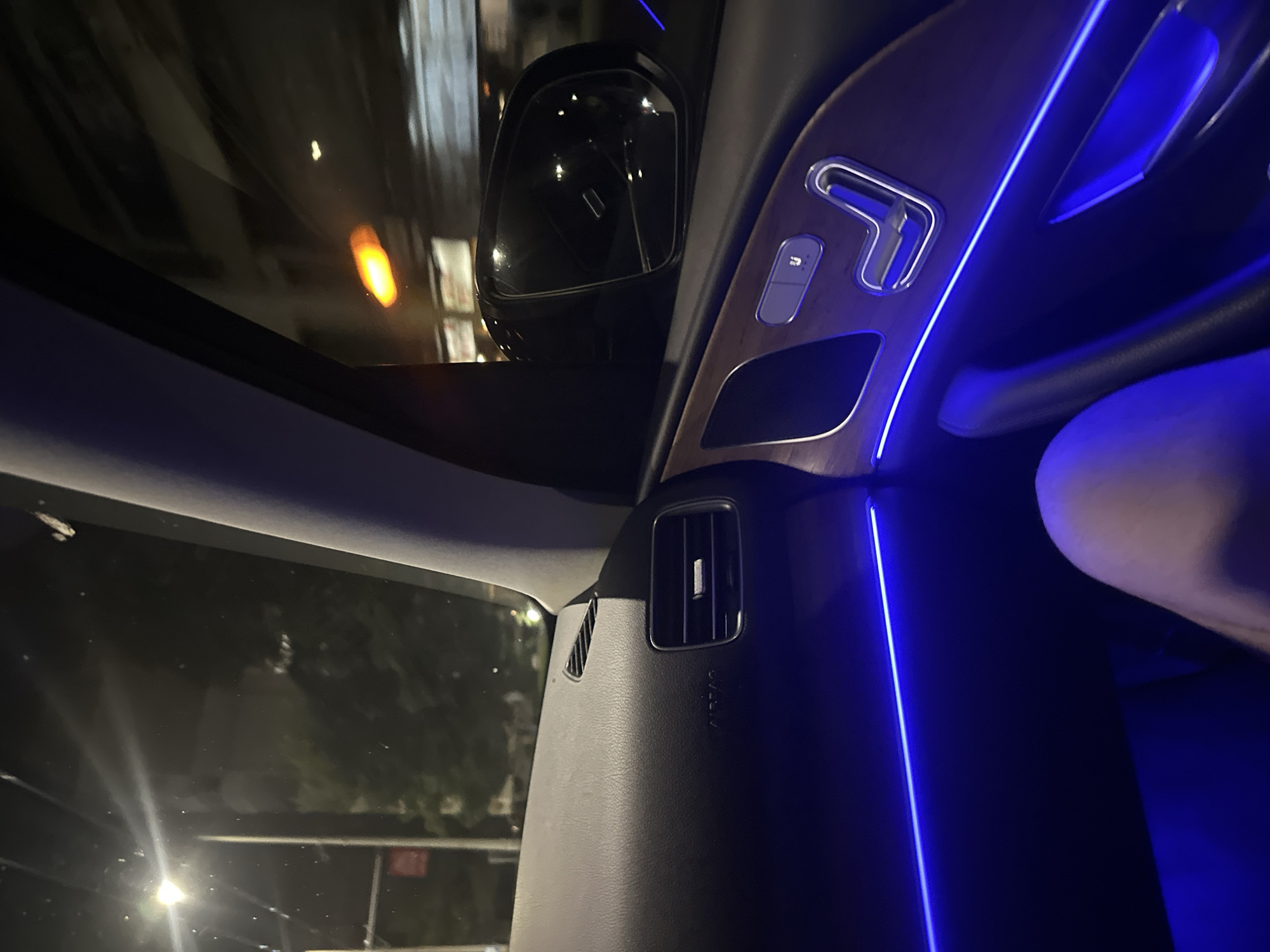 The image captures the interior of a car at night, with blue ambient lighting illuminating the dashboard and door panel. The view is from the perspective of a passenger, looking towards the front and side of the car. Outside the car, streetlights and building lights are visible, suggesting an urban environment. The car's dashboard features an air vent and the word "AIRBAG" is visible. The side door panel has wooden accents and controls for seat adjustment, also highlighted by the blue lighting. The reflection in the side mirror shows lights from passing vehicles or buildings. No specific location in Ruse, Bulgaria is identifiable from the visual cues in the image.