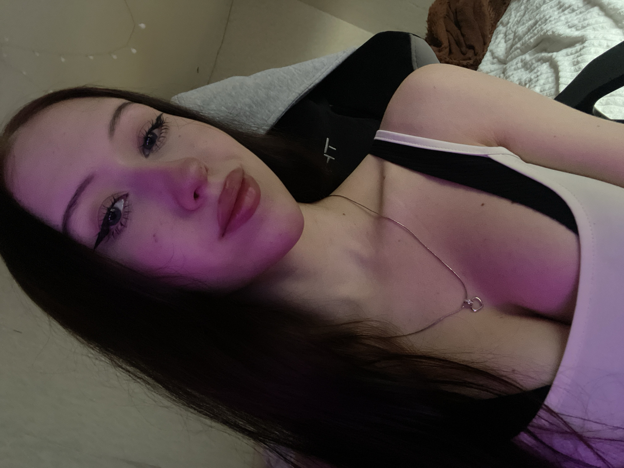 The image is a selfie of a young woman with long, dark hair. She is wearing a white tank top and a black bra. A silver necklace with a heart-shaped pendant is visible around her neck. She has dramatic winged eyeliner and full lips. The lighting in the image is dim with a pinkish hue, possibly from ambient lighting. The background is blurred but appears to be an indoor setting, possibly a bedroom, with a white comforter and what looks like a gaming chair with a grey hood. There are string lights visible in the upper right corner. The scene does not contain any specific landmarks or features that would indicate its location within Votkinsk, Russia.