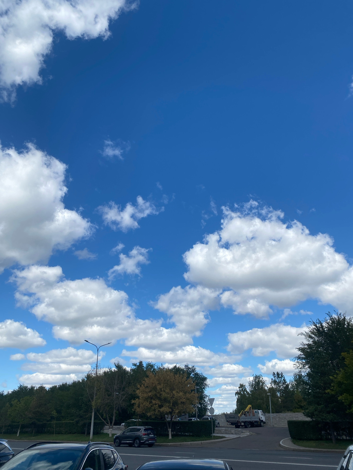 A bright, sunny day in Astana, Kazakhstan, is depicted in this photograph. The sky is a vivid blue, punctuated by fluffy white clouds scattered across the expanse. Below, a line of trees forms a dense green backdrop, with a few trees displaying autumn colors. In the foreground, a road is visible, with cars parked and driving. A street light stands tall, and a construction truck with a yellow crane is parked near a low wall. The scene suggests a typical day in a suburban or semi-urban area of Astana.