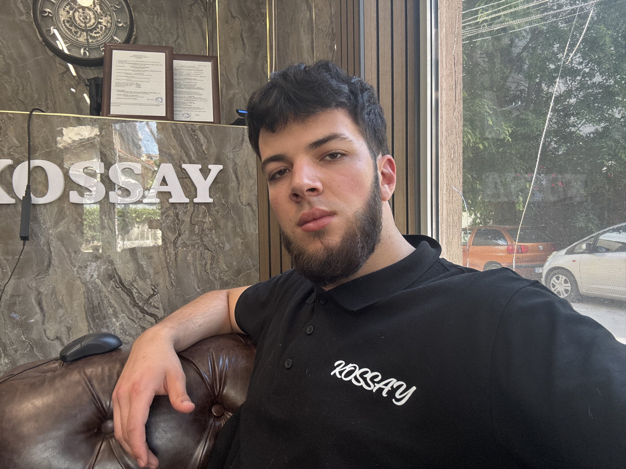 A young man with dark, curly hair and a full beard is seated and looking directly at the camera in what appears to be an indoor reception or office area. He is wearing a black polo shirt with "KOSSAY" written in white cursive text on the left chest. His right arm rests on a brown leather armchair.
In the background, behind the man, is a wall or reception desk clad in a dark, mottled marble-like material. Prominently displayed on this surface are large, white, three-dimensional letters spelling "KOSSAY". Above these letters, on a shelf, are two framed documents, possibly certificates or licenses, and a large, ornate clock with Roman numerals. A computer mouse is visible on the arm of the leather chair beside the man's left hand.
To the right of the man, a large window reveals an outdoor scene during bright daylight. Through a fine mesh window screen, parts of two cars are visible: an orange hatchback on the left and a white car on the right. Green foliage, likely trees, can be seen beyond the vehicles, suggesting a sunny day. The man's expression is neutral and calm. Based on the provided context, this scene is located in Varna, Bulgaria.