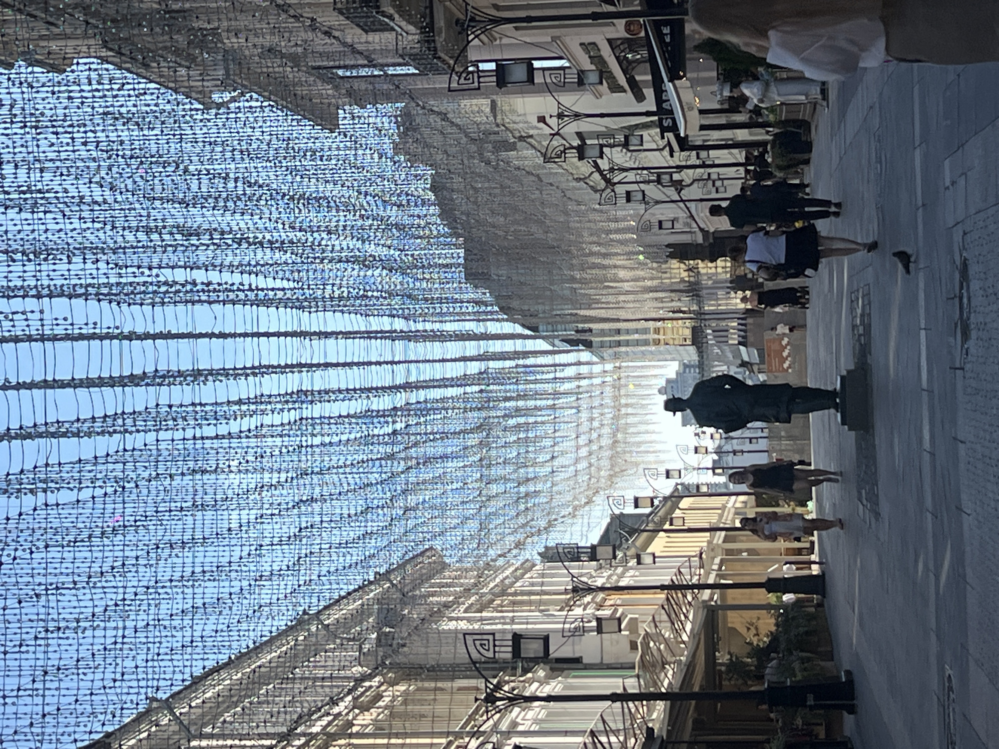 The image depicts a pedestrian street in Moscow, Russia, possibly Nikolskaya Street, adorned with numerous strings of lights hanging overhead. The street is lined with multi-story buildings on both sides, featuring architectural details like balconies and arched windows. Several lampposts with decorative lanterns are visible along the sidewalk.
In the foreground, a statue of a man in a hat stands on a pedestal in the middle of the street. Two women are walking away from the camera on the right side of the street. Further down the street, other people are seen walking, some in groups. The ground is paved with stone slabs. The overall atmosphere suggests a sunny day in a European city.