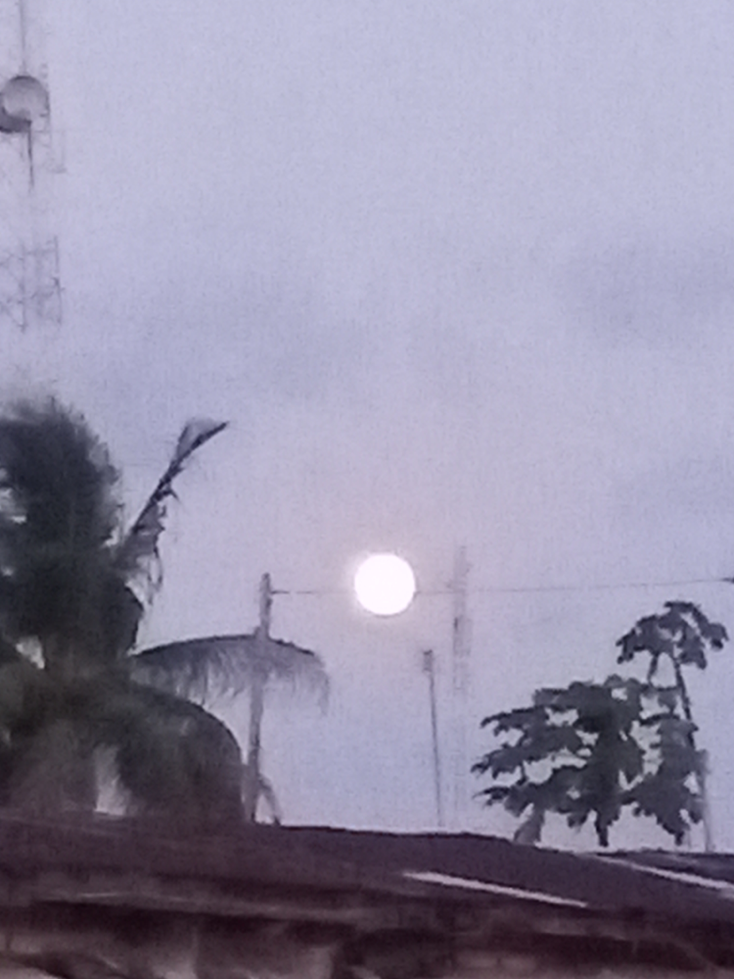 The image depicts an outdoor nocturnal or twilight scene, possibly in Badagry, Nigeria. A dominant, bright circular light source, resembling a full moon or a powerful street lamp, is centered against a soft, uniform light grey-purple sky, suggesting an overcast or hazy atmosphere.
In the foreground, the dark, sloped silhouette of a building's roof, possibly constructed from corrugated metal, spans the lower portion of the frame. Above the roofline, several silhouetted elements are visible. To the left, a large, bushy tree with palm-like fronds extends upwards, partially obscuring a metallic lattice structure that appears to be part of a telecommunication tower or antenna. Another, more slender pole-like structure, possibly another antenna or utility pole, stands to the right of the central light source. On the far right, a different tree with a more dense, branching canopy is silhouetted against the sky.
The scene conveys a sense of stillness and calm, characteristic of evening or early morning hours. No specific activity is taking place, and no people are visible. The soft glow from the central light source and the muted colors create a quiet and slightly mysterious ambiance. There is no visible text in the image.
