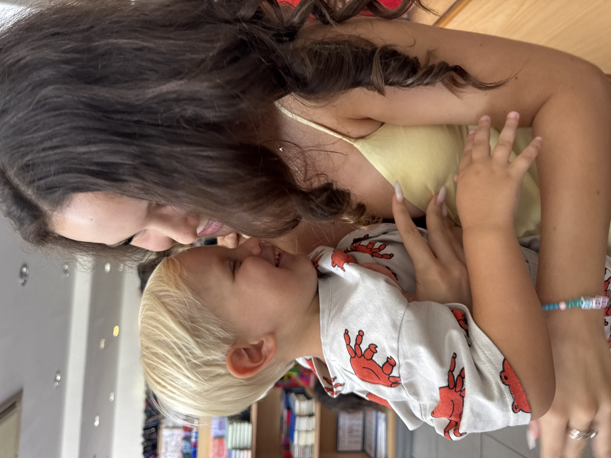 In Agios Athanasios Municipality, Cyprus, a young girl with long, dark, curly hair embraces a blonde-haired young boy. She is wearing a yellow spaghetti strap top, and he is wearing a grey sweatshirt adorned with red crab patterns. The two are close together, both with their eyes closed and smiling. The background appears to be an indoor retail environment, with shelves stocked with various items, possibly books or other merchandise, suggesting a store or library setting. The lighting is bright and even.