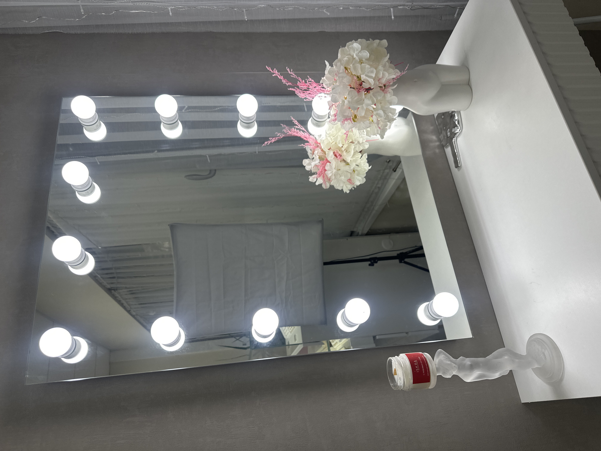 An indoor, eye-level shot depicts a vanity mirror set against a grey wall, adorned with bright, spherical bulbs along its frame. The mirror reflects the room's ceiling, additional lighting, and what appears to be a large white softbox or lighting panel, indicating an illuminated space, possibly a studio or dressing room.
To the right of the mirror, mounted on a white surface, is a decorative arrangement. A white, minimalist bust or torso sculpture, headless and armless, holds a bouquet of artificial flowers. These flowers are predominantly white with delicate pink accents. Below this, a frosted glass sculpture of a reclining female figure extends from the white surface, holding a small, clear glass jar with a red label. The word "ISKRA" is clearly visible on the label.
The composition highlights the bright, artificial lighting from the vanity bulbs and the reflective qualities of the mirror, creating a clean and modern aesthetic. The items are arranged in a decorative fashion, suggesting a well-curated personal or professional space in Sochi, Russia.