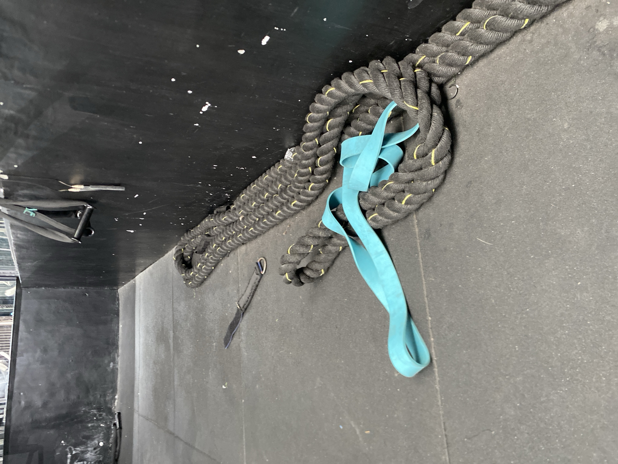 In a gym in North Kuta, Indonesia, a thick, black battle rope with yellow accents lies coiled on a dark gray rubber floor. A bright teal resistance band is looped around a section of the rope. To the left of the rope, a black strap with a metal D-ring lies on the floor. The background consists of a black wall, showing signs of wear and white paint splatters, and a glimpse of a mirrored surface or window at the top.