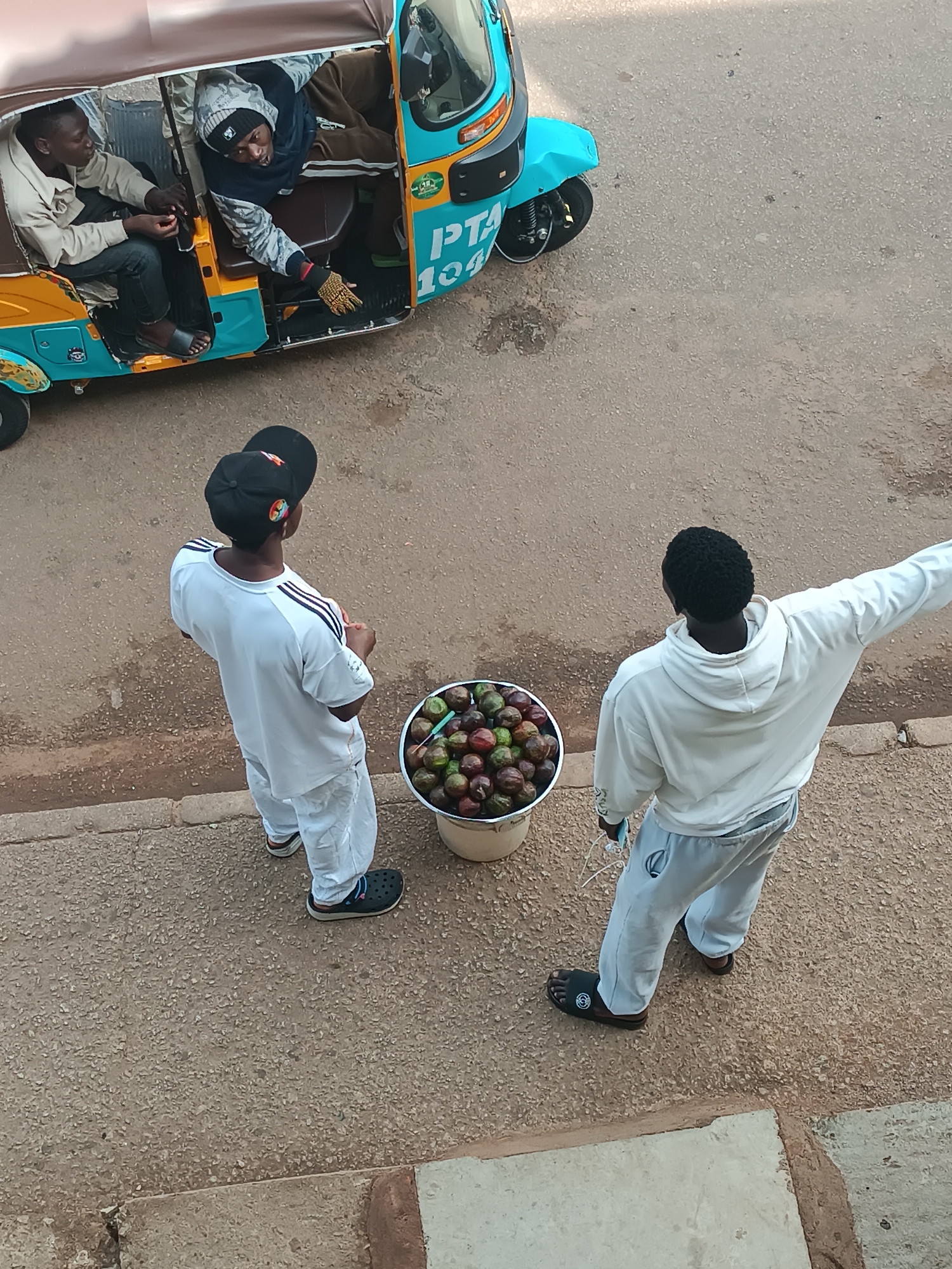 The image captures an outdoor street scene in Bukuru, Nigeria, depicting an interaction involving fruit vendors and passengers in a three-wheeled vehicle.
In the lower half of the frame, two men stand on a paved sidewalk. Between them is a large white bucket filled with small, round fruits, possibly African plums, varying in color from red to green/purple, with small sticks inserted into several. The man on the left wears a white t-shirt with black stripes on the shoulders, white pants, a black baseball cap worn backward, and black crocs. He is seen from behind, looking towards the road. The man on the right, also viewed from the back, wears a white hooded sweatshirt, light grey sweatpants, and black sandals. His right arm is extended outwards, possibly gesturing or pointing.
On the asphalt road next to the sidewalk, a blue and brown/tan auto-rickshaw, or tuk-tuk, is parked. Two men are seated inside. The man on the left, wearing a light-colored jacket, looks towards the fruit bucket. The man on the right, dressed in a dark hoodie with patterns, leans out of the vehicle. His right arm is extended towards the bucket of fruit, and he wears a patterned glove on that hand, suggesting he is reaching to pick fruit or complete a transaction.
Visible text on the side of the auto-rickshaw reads "PTA 104". The scene is illuminated by natural daylight, indicating it is daytime, likely morning or late afternoon. The overall impression is that of a casual street market exchange.