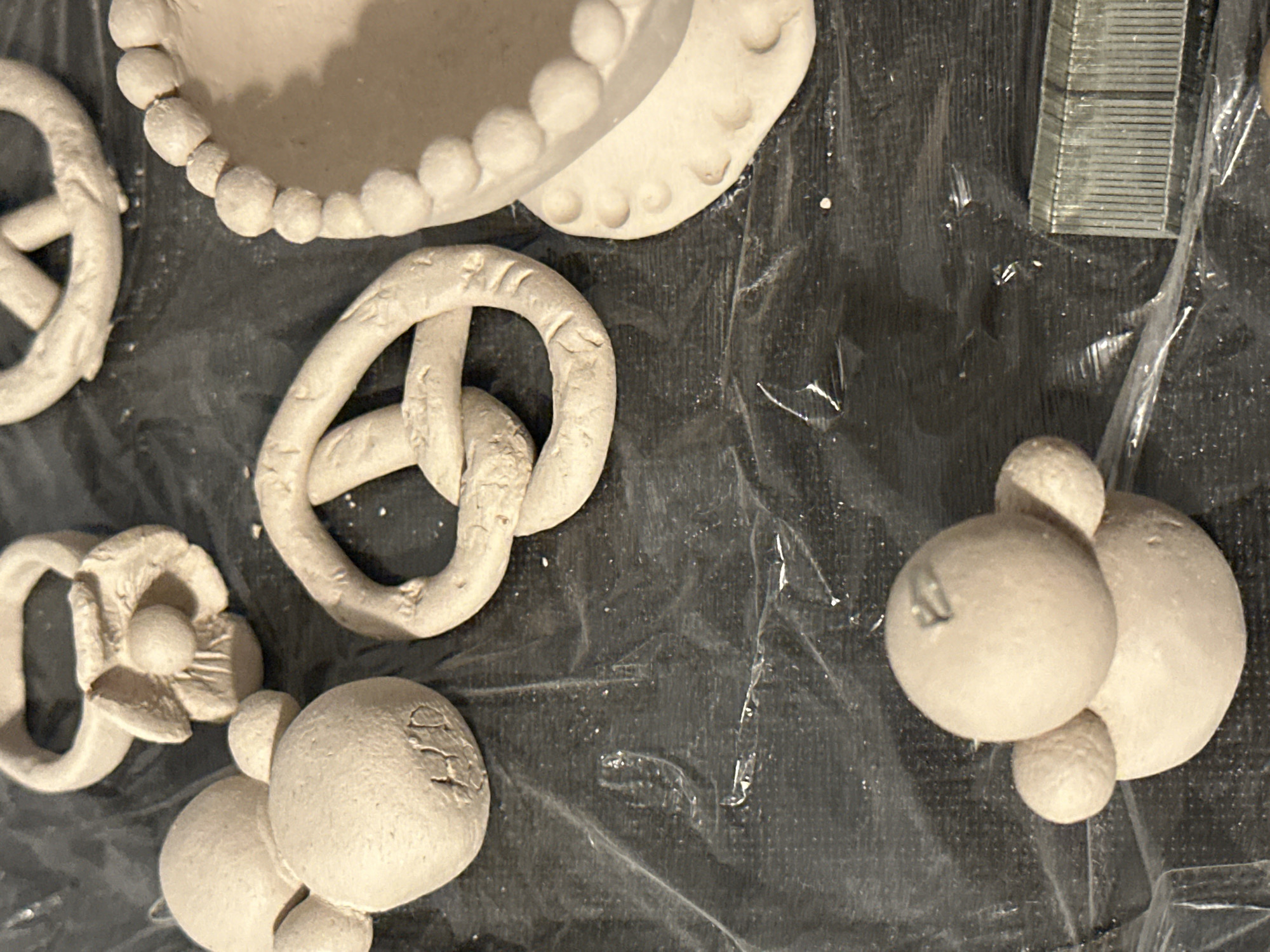 The image shows a collection of unfired clay objects laid out on a black surface covered with plastic sheeting. The objects appear to be handmade crafts, possibly intended for firing in a kiln. In the foreground, a small, round clay figure resembling a bird or a toy is visible. To the upper left, there are two round, ball-like clay shapes, one larger than the other, with a smaller sphere attached to the side of the larger one, resembling a Mickey Mouse figure. Next to this is a clay ring adorned with a flower-like embellishment. In the center of the image, a prominent clay pretzel shape is situated. To the right, a portion of a clay bowl or planter with a decorative beaded rim is visible. The background is a dark, textured surface. The overall scene suggests a crafting activity. There is no identifiable information about the location in Nevinnomyssk, Russia, as the image does not contain any geographical markers or context related to that city.