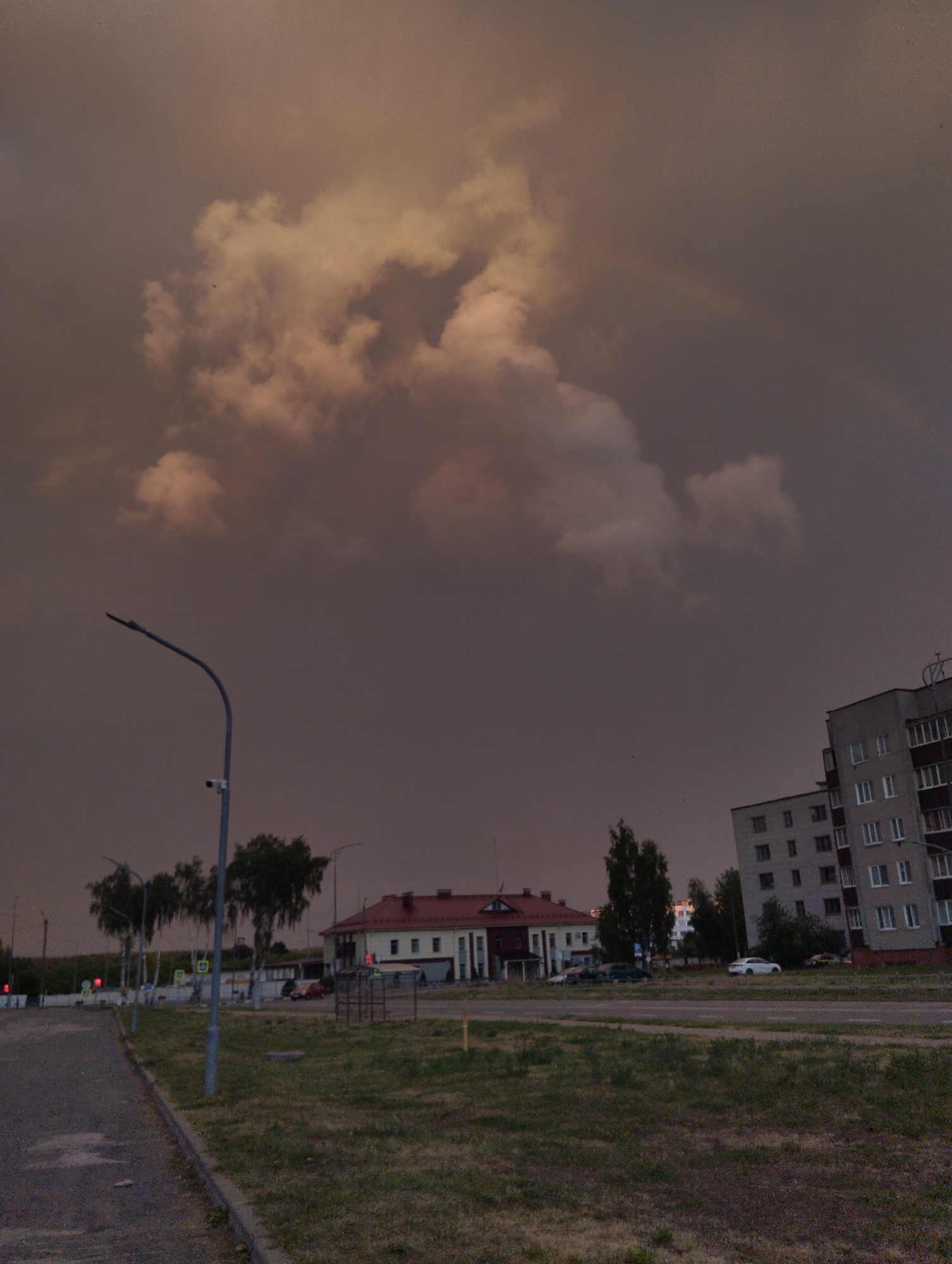 The image depicts an overcast sky with prominent, large, light-colored clouds that appear to be illuminated from behind. A faint rainbow is visible in the upper right quadrant of the sky. In the mid-ground, a multi-story apartment building with multiple windows is situated on the right side. To the left of the apartment building, a low-rise building with a red roof and light-colored facade is visible. In front of these buildings is a paved road with several cars parked on it and a few trees lining the street. A lamppost with a security camera attached is positioned on the left side of the image, near a sidewalk. The foreground consists of a paved pathway or road on the left and a grassy area on the right. The overall lighting suggests either early morning or late evening conditions, with a somber mood due to the dark clouds.
