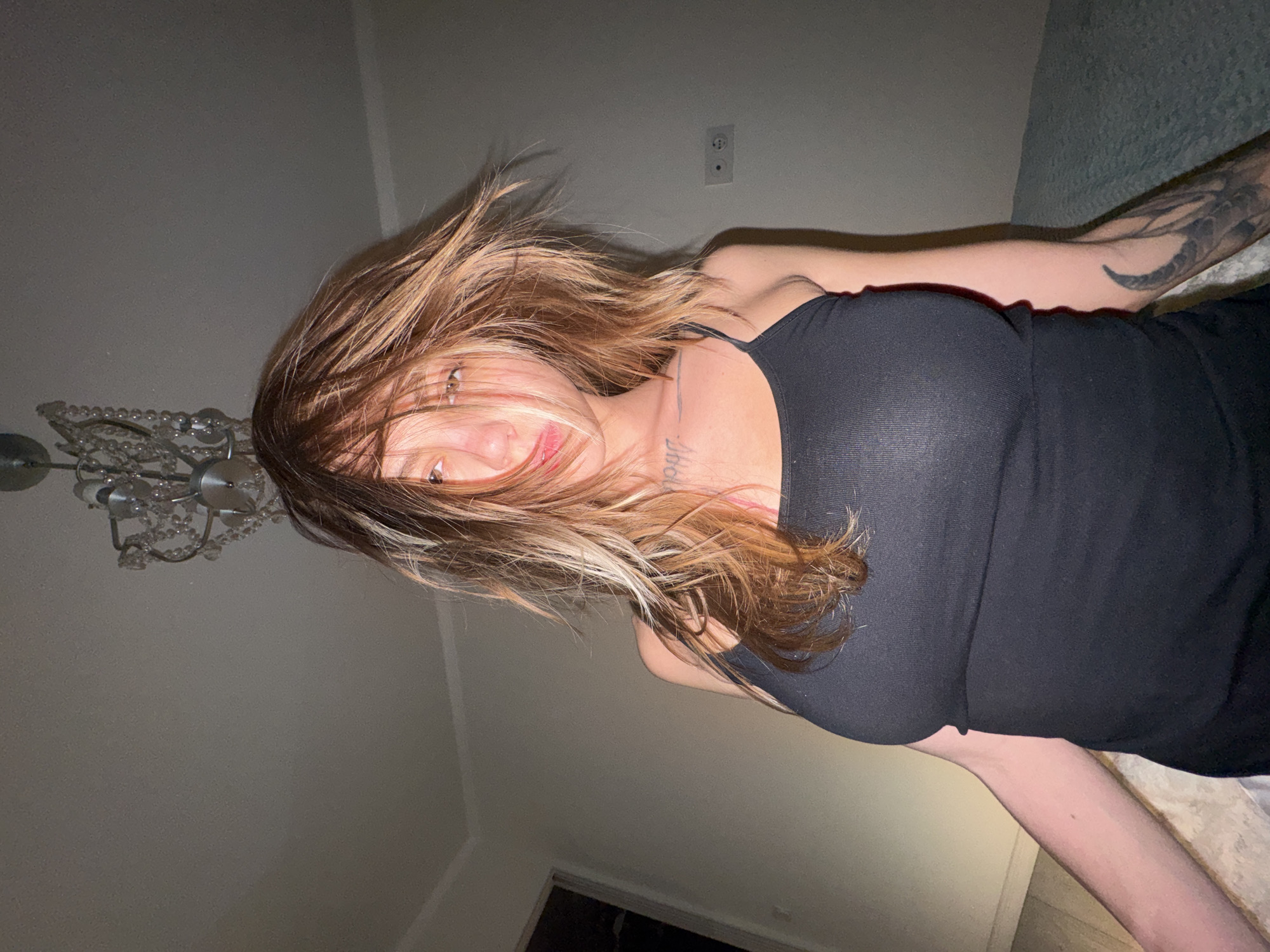 The media file shows a person lying down, possibly on a bed, in what appears to be a dimly lit room. The subject has long, layered brown and blonde hair that is swept across their face, partially obscuring their features. Their eyes are visible and appear to be looking towards the camera. They are wearing a black, sleeveless top. A tattoo is visible on their arm, and some faint markings are visible on their neck, which could be text or other tattoos. To the left of the subject, a chandelier with clear beads is partially visible. The background consists of a plain wall, and to the right, a textured surface, possibly a headboard or wall covering. The lighting suggests it is nighttime or the room is not well-lit. There are no clear indicators of the specific location within Moscow, Russia, other than the provided context. The overall mood seems casual and intimate.