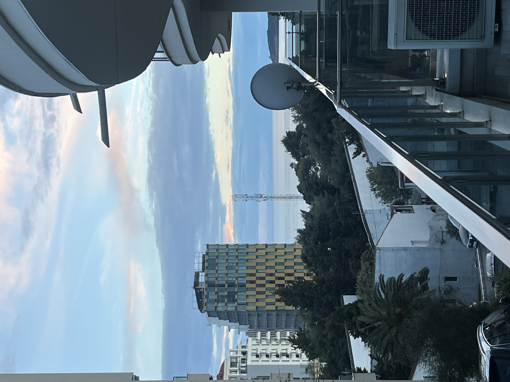 The image depicts an elevated view of Boreti, Montenegro, captured during twilight or dawn. In the immediate foreground, to the right, is a section of a modern balcony featuring a glass and metal railing. A white outdoor air conditioning unit rests on the balcony floor, and a grey parabolic satellite dish is mounted on the railing, oriented towards the sea. Curved white architectural elements of an adjacent or the same building are visible above the balcony.
The midground features a cluster of buildings and dense vegetation. A prominent, multi-story building with a distinctive yellow and dark checkered facade stands centrally, appearing to be under construction or newly completed, with exposed structural elements at its roofline. To its left, other multi-story white residential buildings with balconies are visible. Below these, smaller, older white houses with dark roofs are interspersed among numerous dark green trees, including a prominent palm tree. Portions of parked vehicles, including a dark SUV, are discernible on a street or driveway.
The background reveals the calm, blue-grey surface of the Adriatic Sea. A tall, lattice-structure cellular communication tower is situated on land between the buildings and the coastline. Further across the water, a distant landmass or small island with a faint white structure is visible on the right horizon. The sky overhead exhibits a gradient of light blue, soft pink, and white clouds. No individuals or specific activities are depicted.