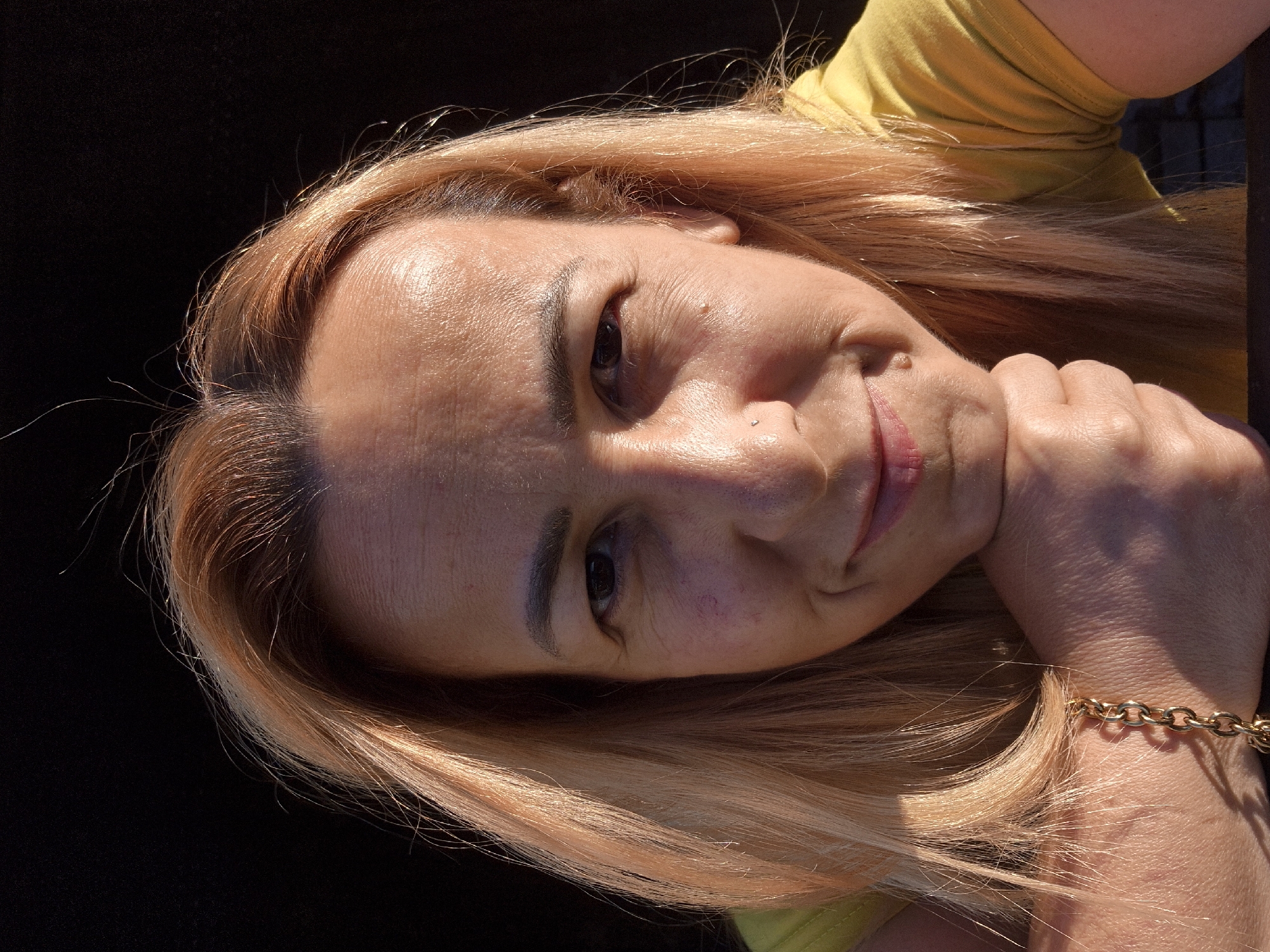 A medium shot shows a woman with blonde hair, a yellow shirt, and a gold bracelet. She is resting her chin on her hand, with her elbow on a dark surface. The woman has a mole on her nose and a small piercing above her lip. The background is dark and out of focus, suggesting an outdoor setting in low light conditions. The woman's skin is illuminated by warm, directional lighting, casting shadows on her face and arm.