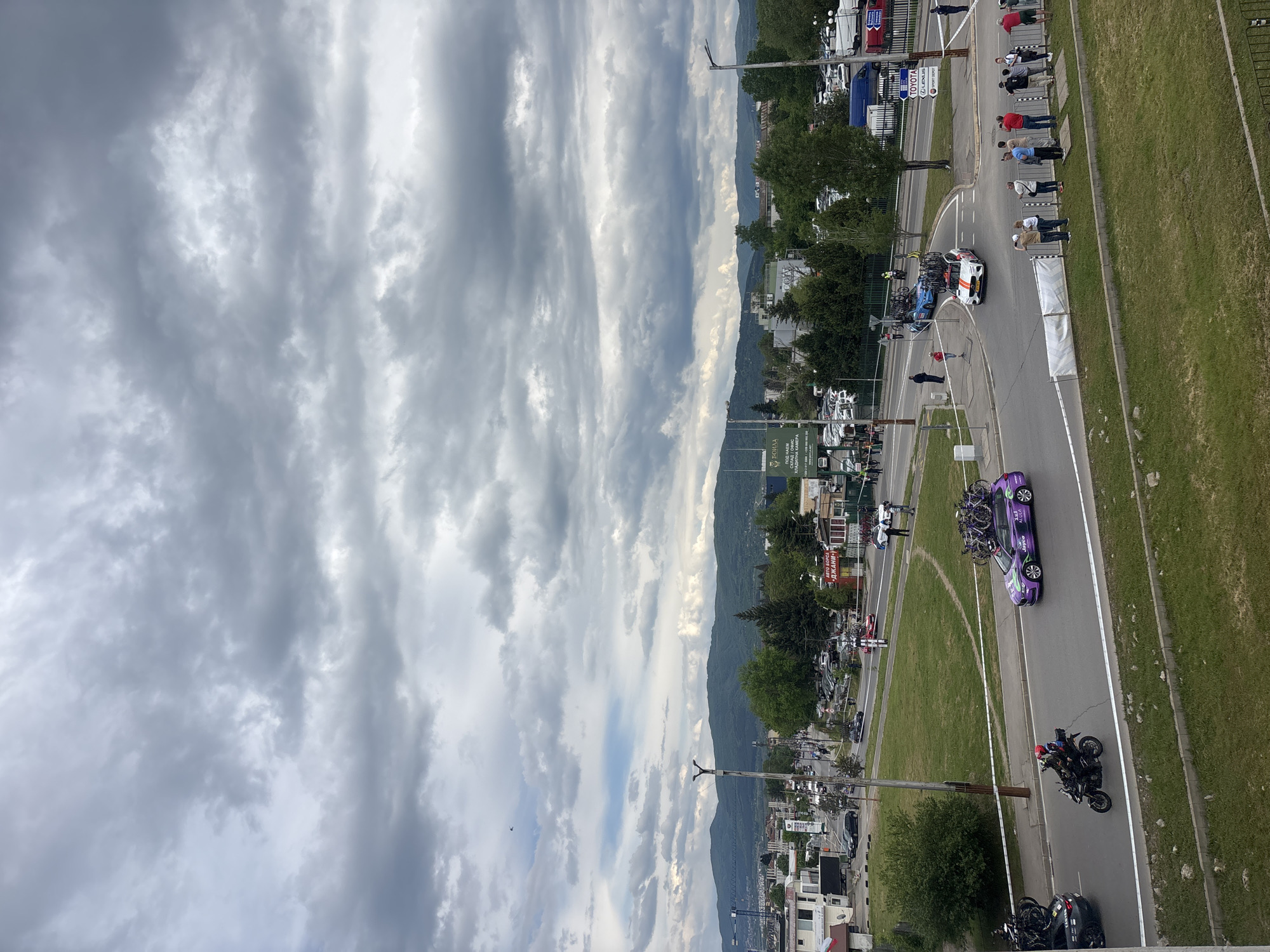 A high-angle, outdoor shot reveals a street scene in what appears to be an urban or suburban area. A street runs horizontally across the lower part of the image, with vehicles and motorcycles visible. A purple car with bicycles strapped to its roof is moving along the road. People are standing on the side of the road, some of whom are behind barriers. The buildings in the background suggest a residential or mixed-use environment, transitioning to trees and greenery. Mountains form the backdrop, and the sky is filled with dramatic, cloudy formations. The city is of Unknown, Unknown.