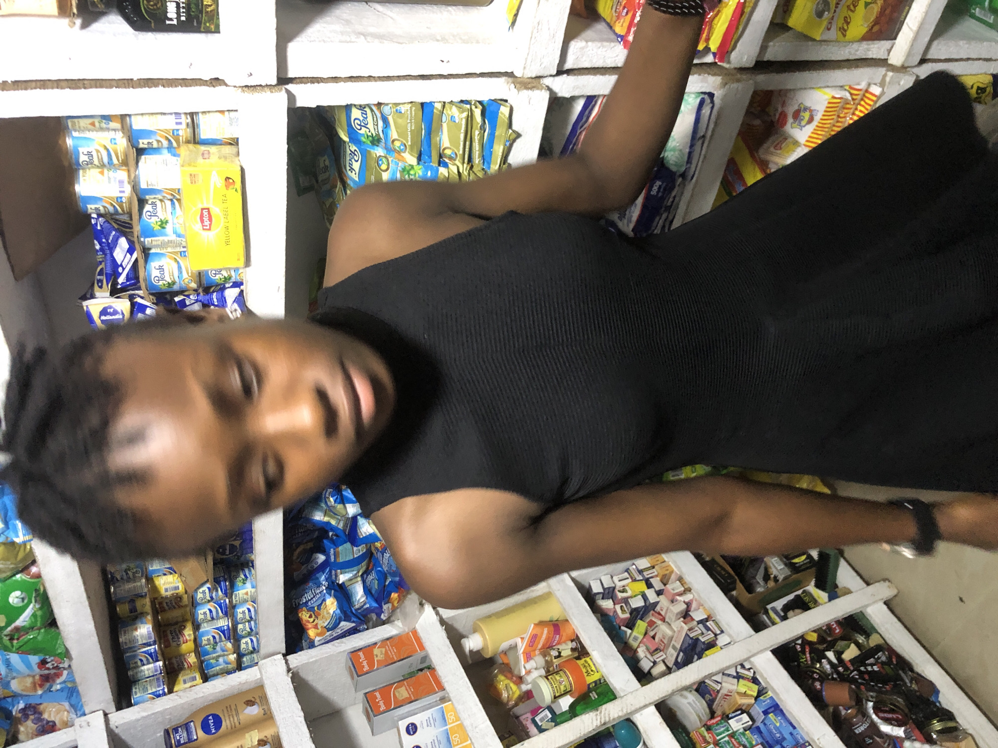 In Ado, Nigeria, a young woman with dark skin and dark braided hair stands in a retail store, her body angled towards the left. She is wearing a black sleeveless dress with a textured fabric. The store shelves behind her are filled with various products, including canned milk, tea boxes, lotions, and packets of food items. To her left, on the shelves, are items such as Nivea cream and other packaged goods. To her right, more shelves are stocked with more packaged food items. The lighting in the store is moderate, illuminating the products and the woman.