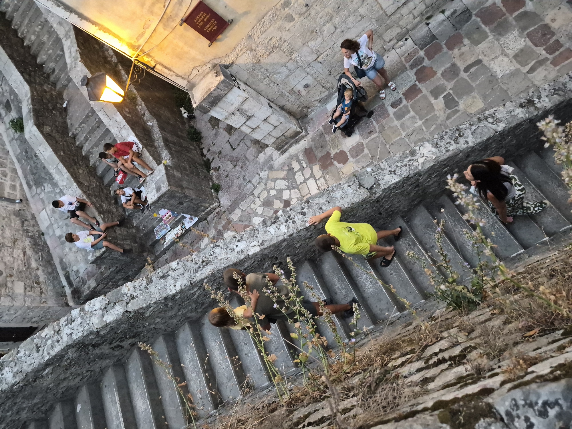 The image shows a view looking down a steep, stone staircase in the old city of Kotor, Montenegro. Several people are present on the stairs and surrounding areas.
In the upper section of the image, several young boys are seated on the steps. One boy in a white t-shirt and blue shorts is sitting on a mid-level step. Further up, another boy in a white t-shirt and dark shorts is also seated. Two more boys are seated on a higher landing, one in a red t-shirt and the other in a grey t-shirt. Scattered on the steps near them are various items, possibly for sale, resembling small trinkets or jewelry.
Descending the main staircase on the left, a woman in a yellow t-shirt is walking downwards, holding the hand of a child on her back. A second woman, with dark hair, is also descending the stairs below them, holding a child on her hip.
In the lower right portion of the image, a woman is standing next to a stroller containing a young child. She is looking slightly upwards and to her left. Another woman with long dark hair is standing on a lower step, looking downwards.
The architecture consists of rough-hewn stone walls and cobblestone paving. A vintage-style street lamp is visible on the right, casting a warm glow. A red banner with some text is affixed to the wall near the lamp. A small black and white cat is visible on the cobblestone ground to the left of the street lamp. The stairs are flanked by stone walls and some dried vegetation. The overall scene depicts a lively, historic urban environment with people navigating the iconic steep streets of Kotor.