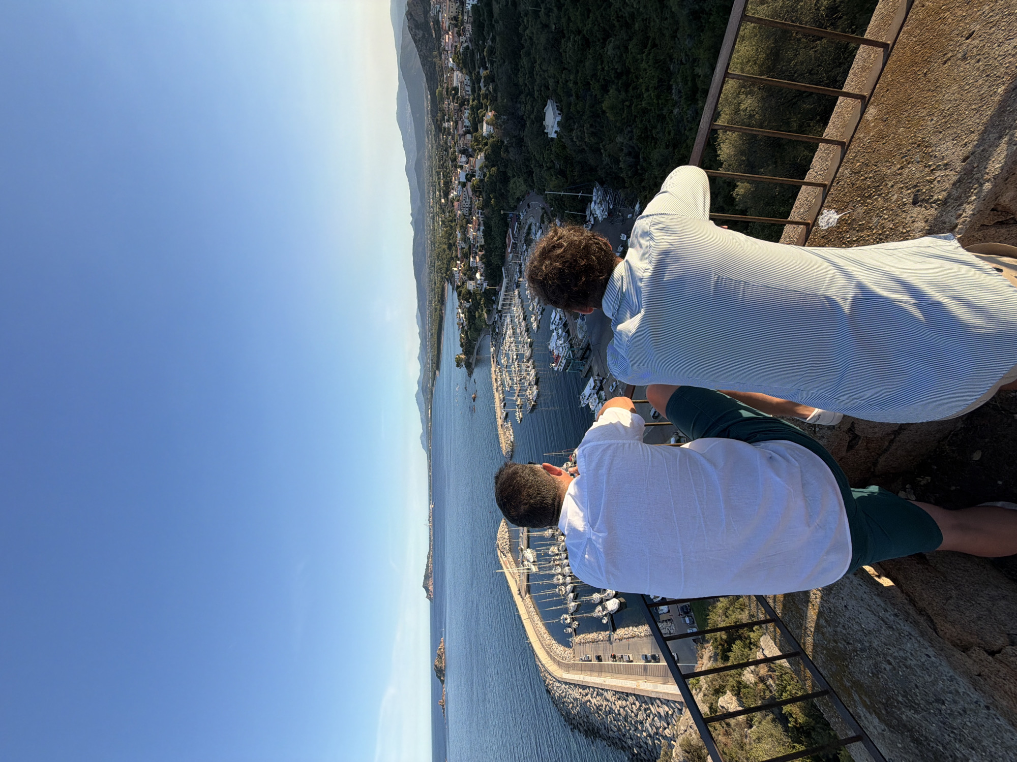 Two men, viewed from behind, stand on a stone ledge overlooking a scenic coastal town and harbor. The man on the left wears a white, short-sleeved button-down shirt and green shorts. The man on the right wears a light blue and white striped shirt. They are both leaning on a metal railing, looking out at the view. The harbor is filled with numerous sailboats. Beyond the harbor, the town is nestled amongst green hills, with buildings featuring red-tiled roofs. The sea stretches out to the horizon, with islands visible in the distance under a clear blue sky. The scene depicts a picturesque view of Santa Maria Navarrese, Italy, at golden hour.