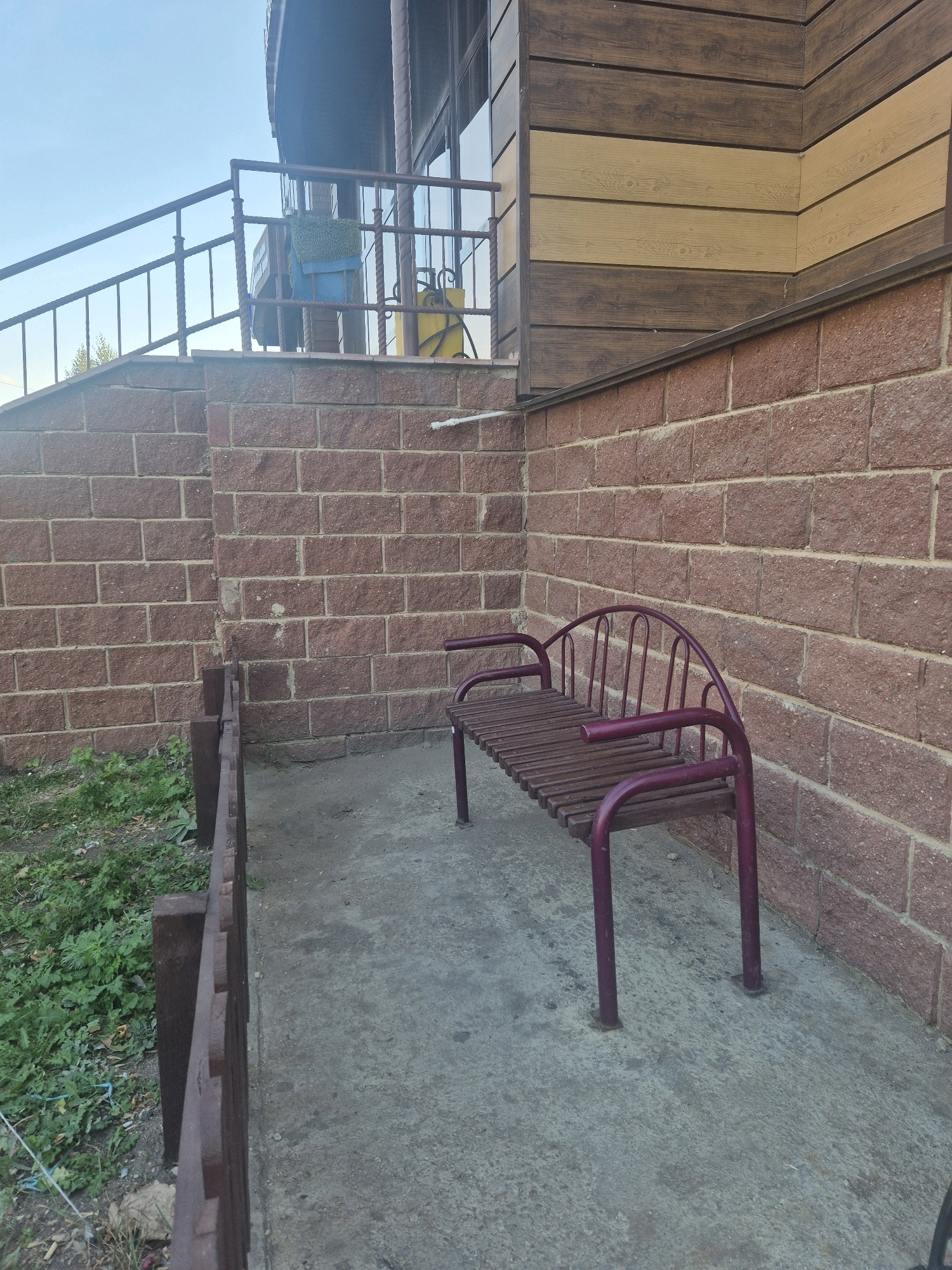 An outdoor scene features a purple metal bench with a wooden slatted seat and backrest. The bench is positioned on a concrete pad next to a reddish-brown brick wall. To the left of the bench, a low wooden fence separates the concrete area from a patch of green grass and weeds. In the background, a portion of a building is visible, with its upper section clad in horizontal wood siding, alternating between dark brown and light tan. A staircase with a metal railing leads up to the building, and a blue object and a green textile are draped over the railing. The sky above is a clear, light blue. The image does not contain any specific landmarks or clues to pinpoint its location within Kokshetau, Kazakhstan.