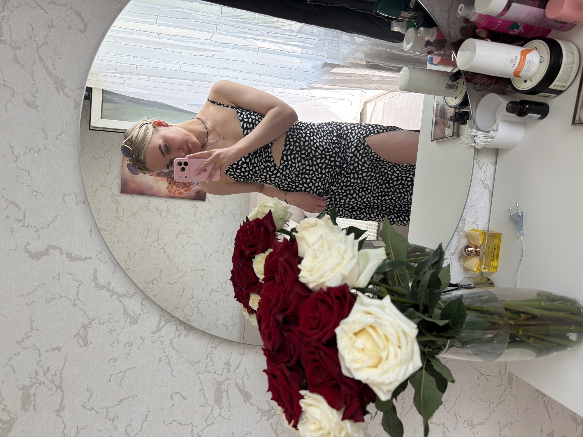 A photograph shows a woman taking a selfie in a mirror. The woman has blonde hair and is wearing sunglasses pushed up on her head. She is wearing a black and white patterned dress. The background includes a window with sheer white curtains. In the foreground, a bouquet of red and white roses is in a vase. On the table, there are assorted cosmetic products and a bottle of perfume. The location is Unknown, Unknown.