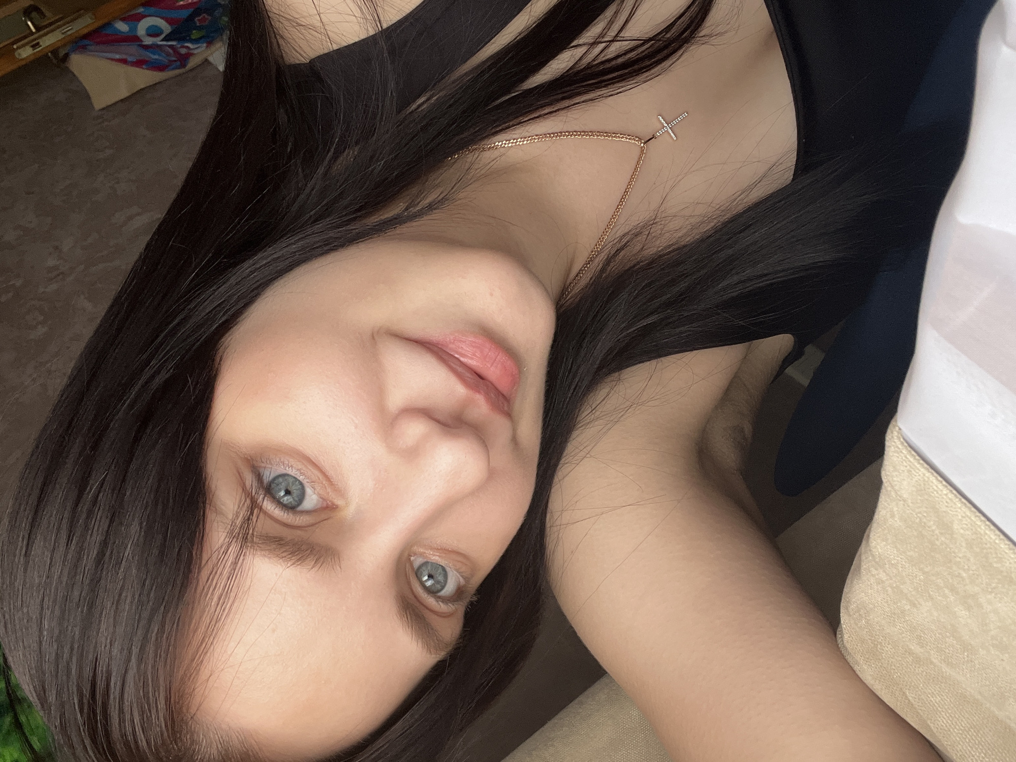 A young woman with long dark hair and blue eyes gazes at the camera. She is wearing a black top and a delicate gold necklace with a cross pendant. She appears to be indoors, possibly in a bedroom, based on the presence of a bed and other furniture in the periphery. The location is Марковский, Russia.