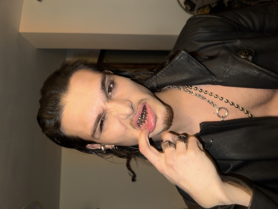 The image presents a medium close-up of a young male individual, estimated to be in his late teens or early twenties, directly facing the camera. He possesses dark, medium-length hair styled away from his face, dark eyes, and pronounced eyebrows. A small goatee and light stubble are visible on his chin and upper lip. The subject exhibits a contorted facial expression, characterized by a wrinkled nose, narrowed eyes, and an open mouth. His left index finger is positioned to pull down his upper lip, intentionally exposing a silver-toned metallic dental grill (grillz) fitted over his upper teeth. The grill appears to be designed with multiple conical or helical protrusions.
The individual is dressed in an open black leather jacket, revealing a bare chest. He wears two distinct silver-toned necklaces: one is a prominent chain featuring a circular ring pendant, and the other is a longer ball chain. On his left hand, he wears at least two rings, one on his ring finger and another on his middle finger. His left ear is adorned with multiple piercings, including a hoop earring, a stud, and a small dangle earring.
The background is indistinct, indicating an indoor environment with plain, light-colored walls, likely beige or off-white. A room corner is visible on the right side of the frame. The lighting is warm and generally uniform. While the geographical context is provided as Sofia, Bulgaria, the specific location within the city cannot be determined from the visual information available in the image.
