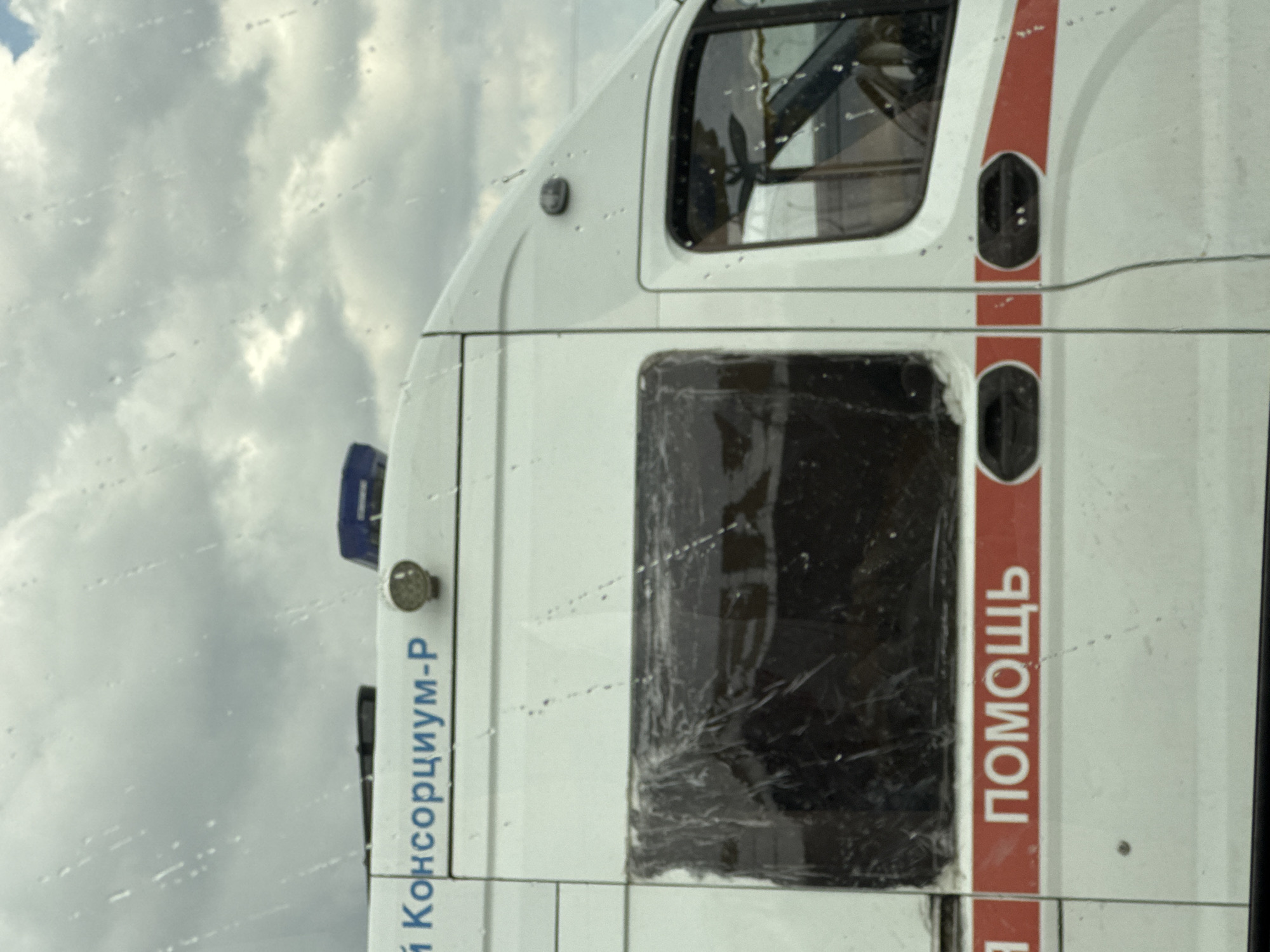 A white ambulance with a red stripe is parked on a street. The ambulance has a black window and some text on its side. The text on the side of the ambulance is in Russian and reads "ПОМОЩЬ" (help) and "Й КОНСОРЦИУМ-Р" (Y Consortium-R). The sky is cloudy, suggesting it may be daytime. The image is taken from a slightly low angle, looking up at the vehicle. There are water droplets on what appears to be a window, possibly indicating rain or a dirty windshield. The overall scene suggests a typical street view of an emergency vehicle in Russia.