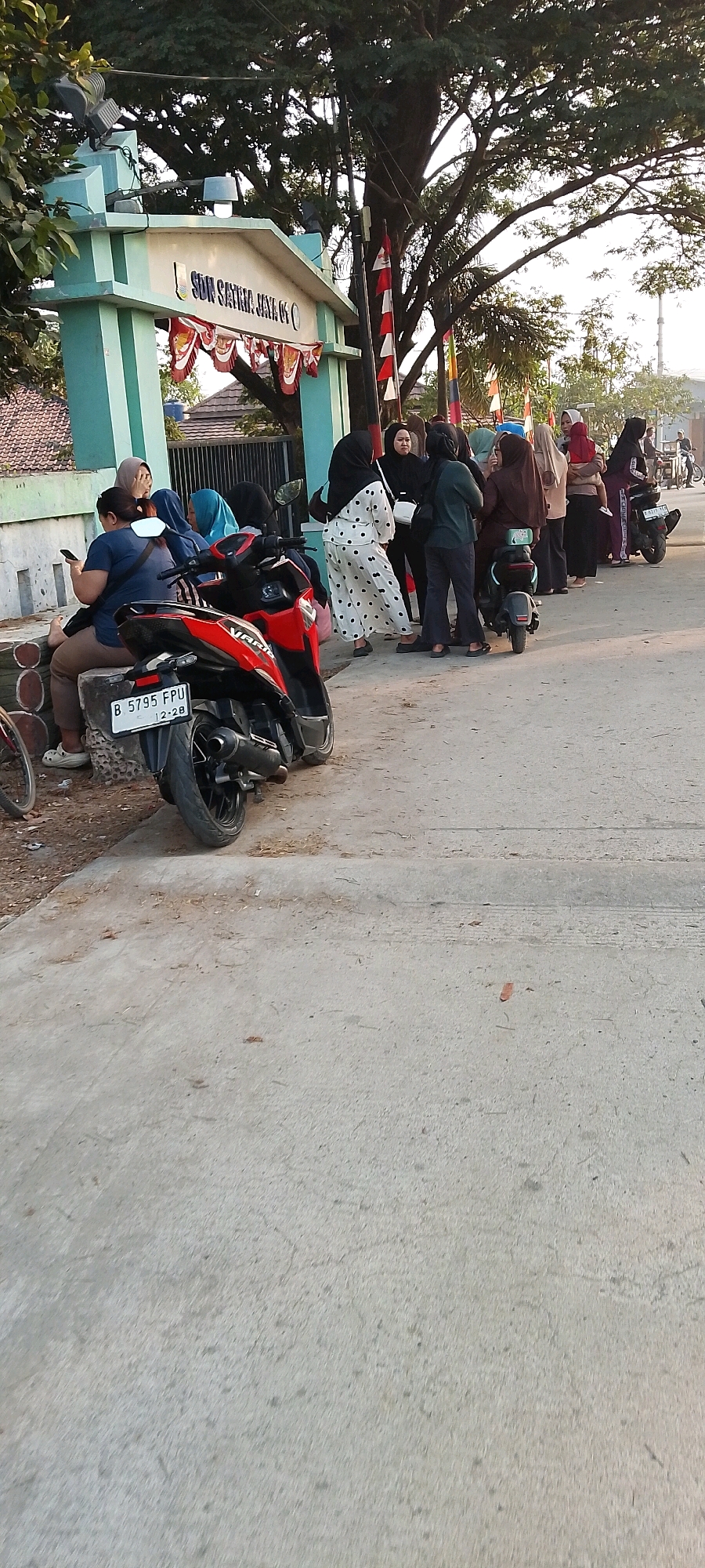 The image depicts a street scene in front of a building with signage that reads "SDN SATRIJAYA JAYA" in Satriajaya, Indonesia. Several individuals, primarily women wearing headscarves, are standing in a line, some appearing to be waiting. A red and black motorcycle is parked in the foreground, partially obscuring a concrete seating area where a person is seated while looking at their phone. Another motorcycle is visible further down the street. The environment appears to be outdoors, with a large tree casting shade over the entrance to the building, and flags decorating the entrance archway. The time of day appears to be late afternoon or early evening, given the lighting conditions.