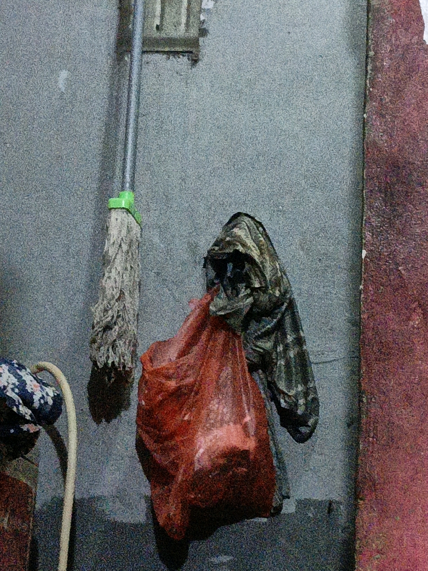 A mop with a green handle and frayed white strings hangs vertically against a grey, textured wall. To the right of the mop, a dark, hooded garment is draped over something unseen, with a translucent red plastic bag hanging in front of it, filled with what appears to be dark, lumpy contents. To the left of the mop, a white curved hose-like object is visible, with a portion of a patterned textile, possibly fabric, attached to its upper end. A reddish-brown textured surface runs vertically along the right edge of the frame. The scene appears to be an outdoor space in West Jakarta, Indonesia, likely an alley or a service area given the utilitarian objects and the rough wall texture.
