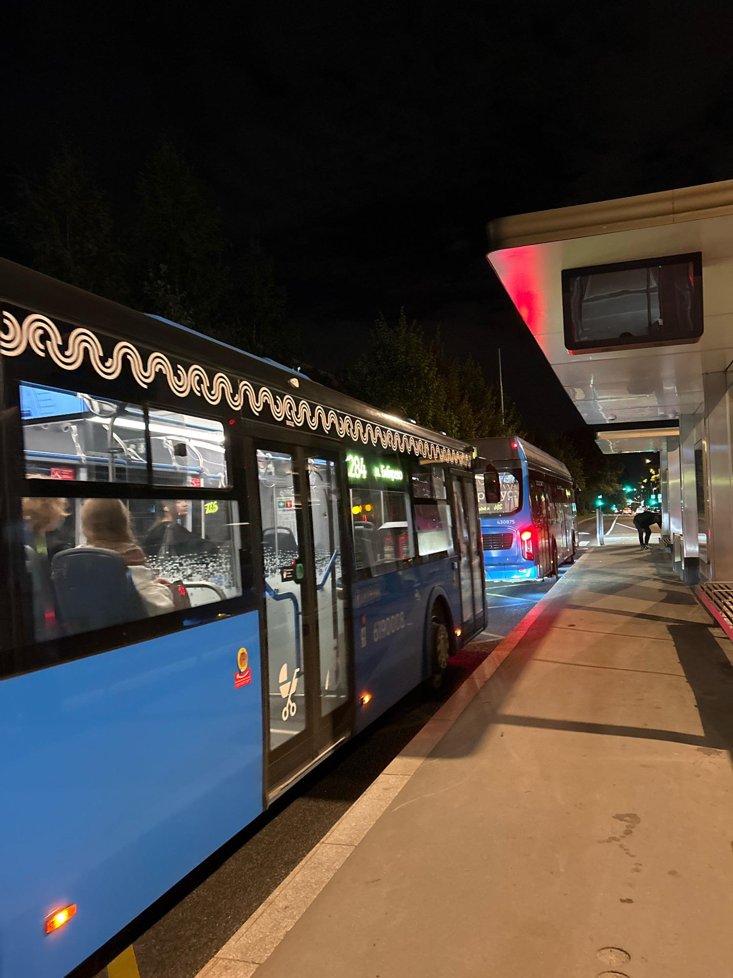 Two blue city buses are parked side-by-side at a bus stop at night. The bus in the foreground has its side facing the viewer, revealing passengers inside. The bus in the background is partially visible, with its rear end closest to the foreground bus. The bus stop appears to be a modern structure with a covered waiting area, illuminated by red and white lights. Trees and darkness are visible in the background, suggesting an urban setting. A person is standing near the back of the bus stop, facing away from the camera, appearing to interact with something on the ground. The overall scene depicts a typical urban transit environment during nighttime hours. This image was taken in Moscow, Russia.
