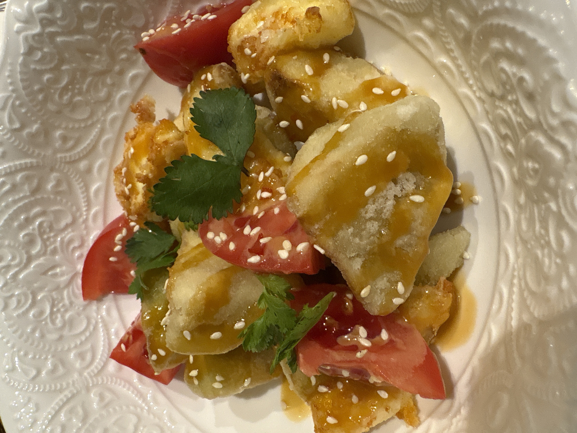 A close-up shot shows a white, ornate bowl filled with a dish that includes pieces of fried food covered in a glaze and sesame seeds, interspersed with wedges of ripe, red tomatoes. Sprigs of fresh green cilantro are also visible, adding a touch of freshness to the dish. The bowl has a patterned rim, suggesting it might be made of porcelain or a similar material. The setting is likely indoors, perhaps a home or restaurant, given the context of the served dish. There is no clear indication in the image that it was taken in Kazan, Russia.