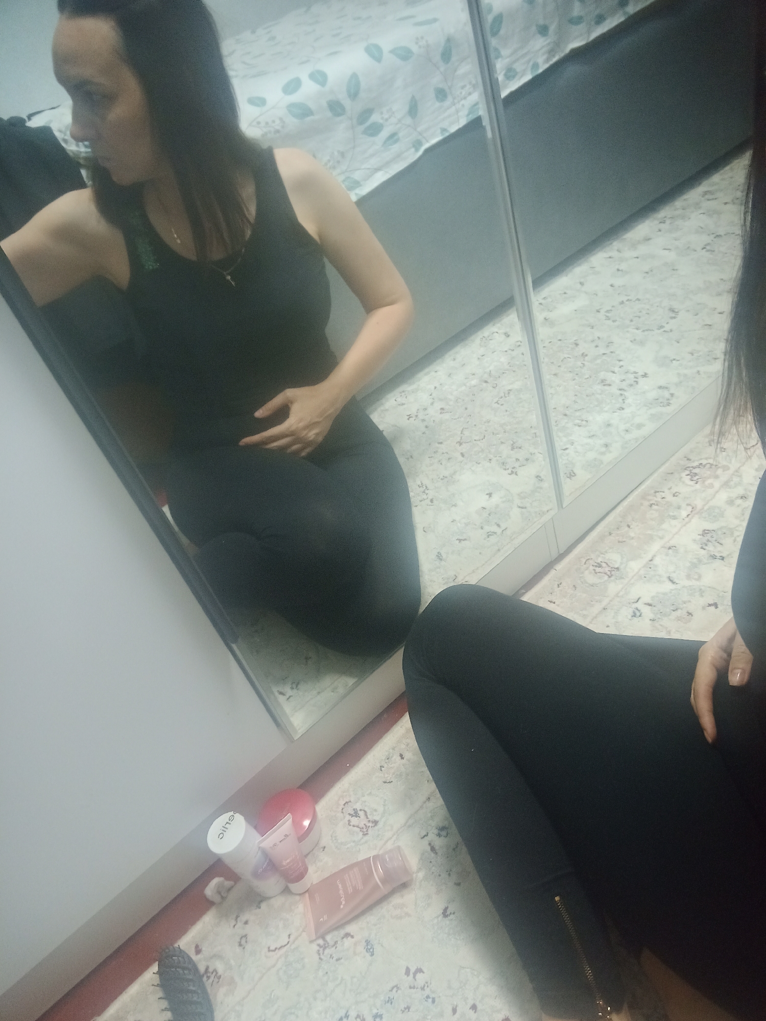 The image shows a woman in black leggings and a black tank top sitting on the floor in front of a mirrored closet. She is looking at her reflection and has one hand on her stomach. There are several cosmetic products scattered on the floor in front of the closet. The room appears to be a bedroom, with a bed and bedding visible in the background. The overall scene is casual and domestic. The location in the city of Osh City, Kyrgyzstan is not evident from the image.