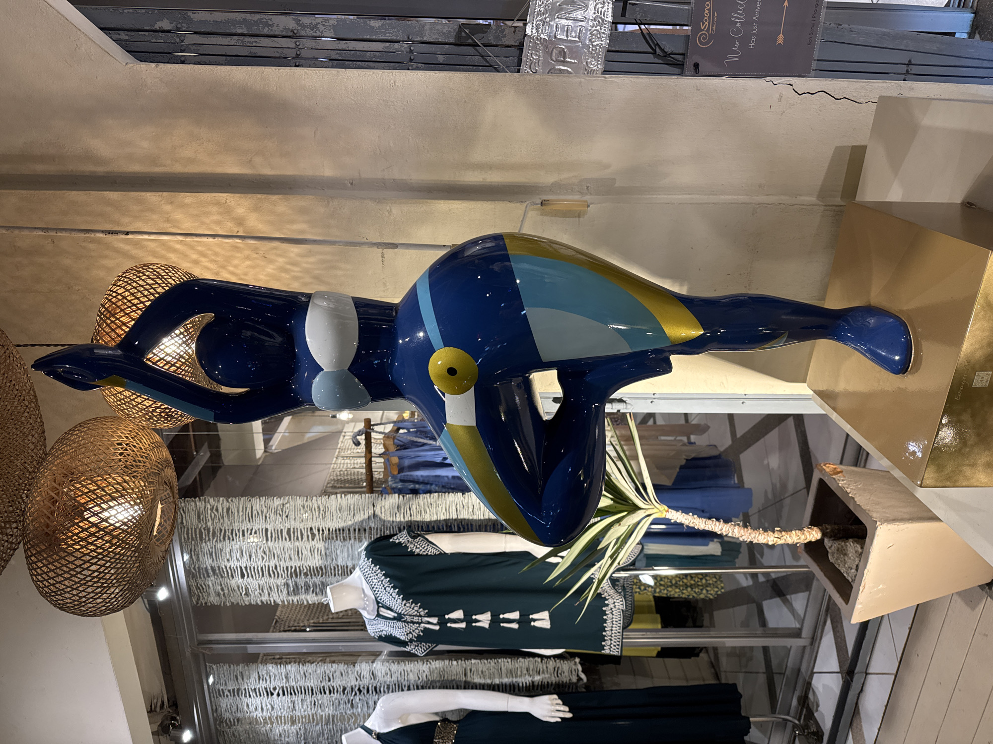 The image depicts the interior of a shop in Baan Bo Phut, Thailand. A blue and gold painted mannequin in a yoga pose is a prominent feature, with a woven lampshade beside it. The mannequin is positioned above clothes displayed on mannequins and racks. A potted plant is visible beside the base of the painted mannequin. An "OPEN" sign and "New Collection" advertisement are visible near the top of the frame.