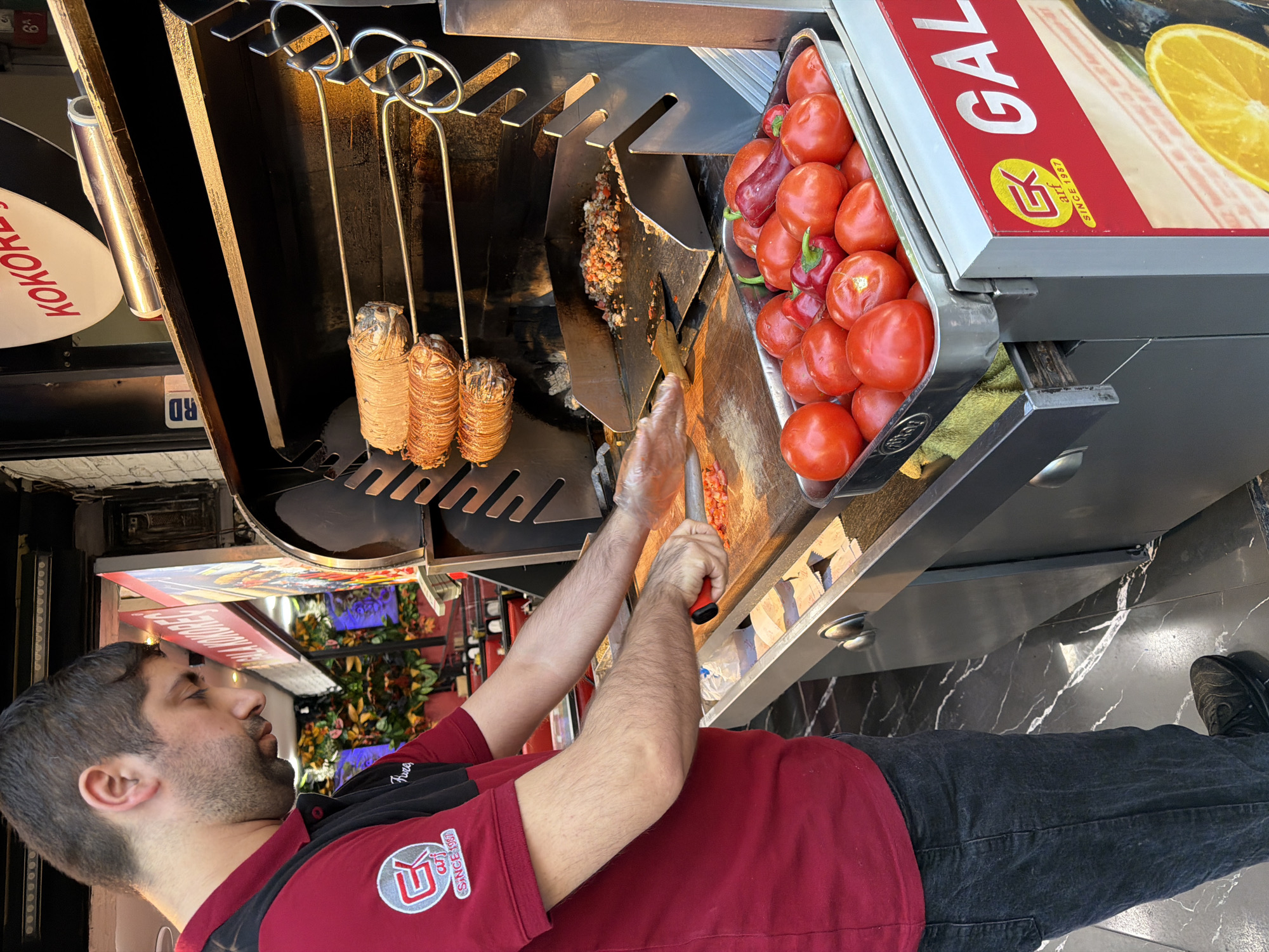 A man in a red and black polo shirt with a circular logo is preparing food in front of a rotating spit of meat, possibly kokoreç, at a street food vendor. He is wearing a red polo shirt with a black panel on the sleeves and shoulder. The logo on his left breast is yellow with a red outline and appears to say "GK" with "SINCE 1987" below it. He is wearing dark denim jeans and black shoes.
The vendor has a large metal grill with the meat rotating on skewers above a hot plate where chopped ingredients are being cooked. In the foreground, a metal tray is filled with ripe red tomatoes and a few red peppers. A cutting board with chopped tomatoes and a red-handled knife are visible on the counter. The vendor is holding a spatula in his right hand, appearing to mix the ingredients on the hot plate.
Behind the vendor, there are signs and decorations, including a red and white sign with Turkish lettering that says "KOKOREÇ". The flooring appears to be black marble with white veins. To the right of the vendor, a red and white sign with "GAL" in large white letters is visible, suggesting the name of the establishment. The overall scene depicts a busy street food environment in the Üsküdar district of Istanbul, Turkey, where this popular dish is commonly served.