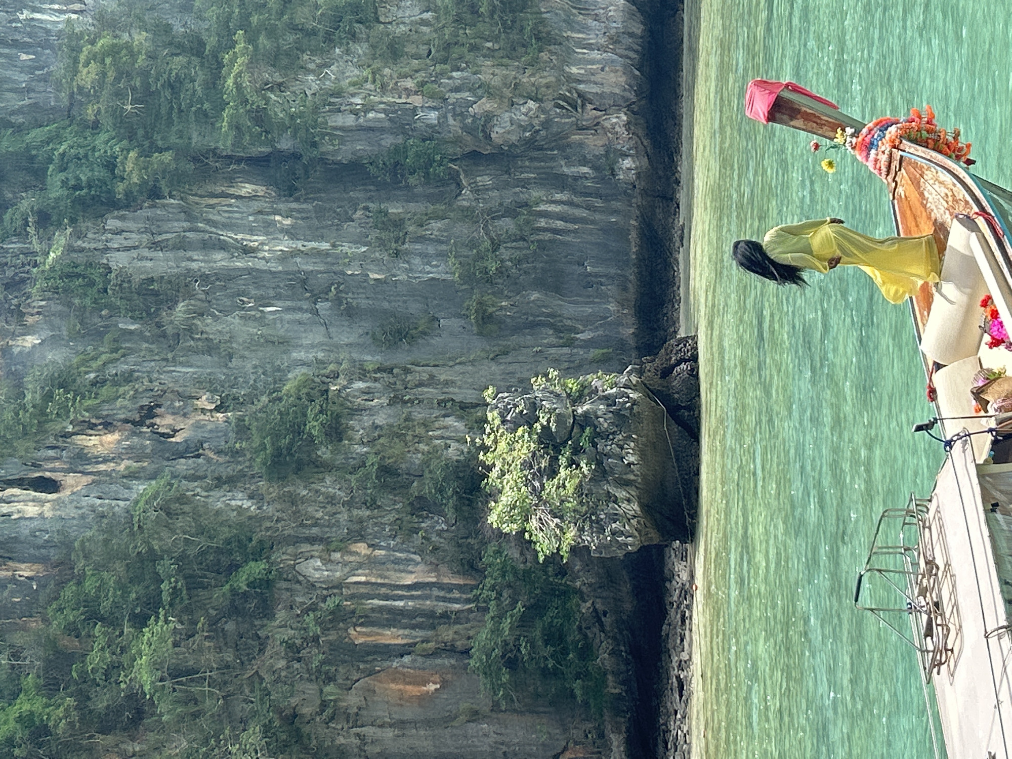 A woman stands on the bow of a longtail boat in Ao Nang, Thailand, facing away from the camera and looking out at the turquoise water. She is wearing a flowing yellow dress that billows in the wind. The boat is decorated with colorful garlands and flowers. In the background, a sheer limestone cliff rises dramatically from the water, covered in lush green vegetation. A small, rocky island topped with trees sits in the foreground, between the cliff and the boat. The water is clear and appears shallow in some areas. The scene is bathed in bright sunlight.
