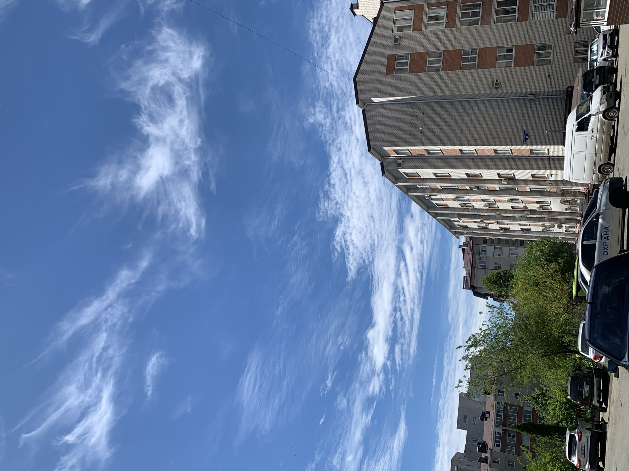 This media file captures an urban street scene in Stavropol, Russia, on a bright, sunny day with a clear blue sky dotted with wispy white clouds. The original image is oriented vertically, rotated 90 degrees clockwise, making the sky appear on the left and the ground with buildings and vehicles on the right.
The dominant subjects are a multi-story building and several parked vehicles. The main building on the right features light grey brickwork with rectangular windows framed by brown or orange accents. Numerous external air conditioning units are visible on its facade. In the background, other residential or commercial buildings are partially visible, interspersed with green trees and bushes, suggesting a well-vegetated urban environment.
Along the bottom and right side of the frame, several cars are parked. A prominent white cargo van is seen, along with a dark SUV and a lighter-colored sedan. There is no discernible activity or event taking place; the scene appears still.
Notable details include the bright daylight, indicating mid-day, and the weather, which is clear with high, scattered clouds. Location cues are present through visible text. On a dark vehicle, likely a security patrol car, the Cyrillic word "ОХРАНА" (OKHRANA), meaning "Security," is clearly visible in white letters. License plates on two vehicles are also partially legible: "к534хс 26" on the dark SUV and "о583тс 26" on the lighter sedan. The "26" is the regional code for Stavropol Krai, confirming the provided location context. No people or emotions are visibly captured in the image.