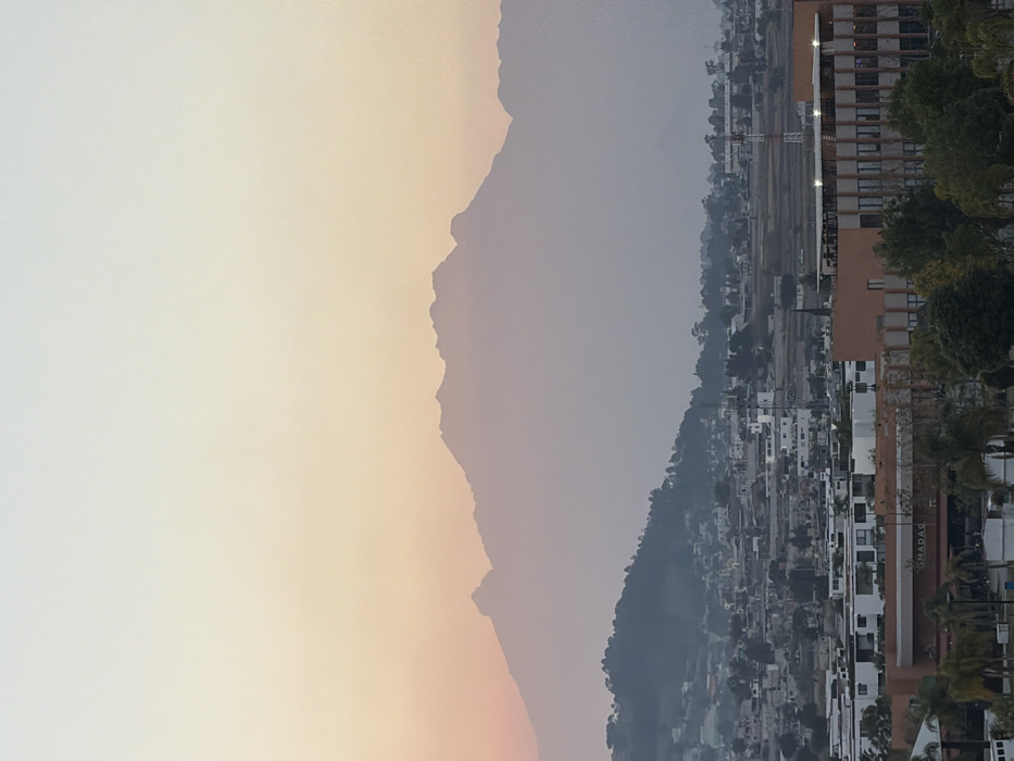 A hazy, low-lit landscape features a mountain range silhouetted against a pale sky transitioning from light orange to gray. A dense urban area lines the right edge, consisting of tightly packed buildings of varying heights, some obscured by trees. A crane is visible amidst an open, flat area within the town. The entire scene is oriented vertically.
